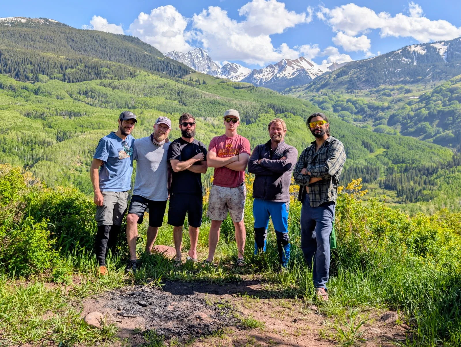 Six men standing outdoors against a backdrop of green mountains and blue sky with clouds, some wearing sunglasses and casual clothing.