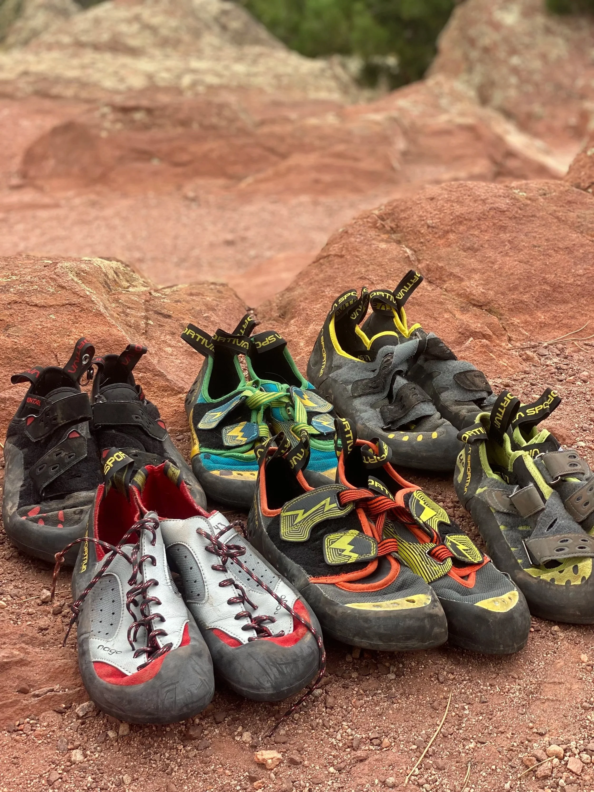 Six pairs of colorful climbing shoes arranged on rocky ground outdoors.