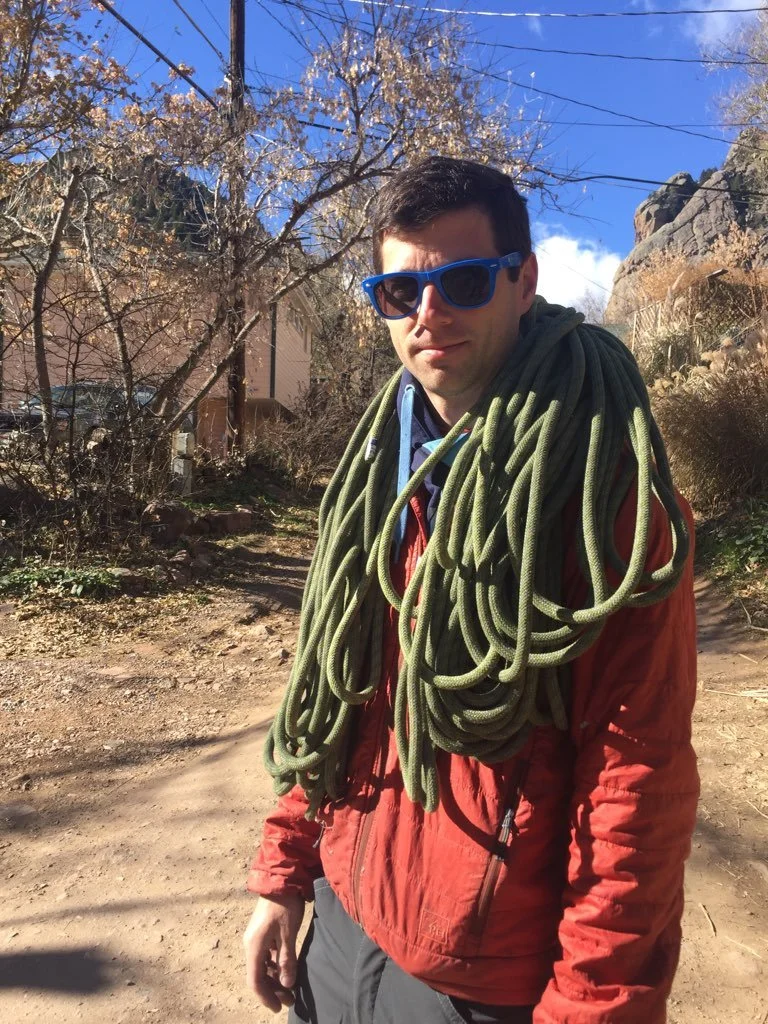 A man wearing sunglasses and a red jacket outdoors on a dirt path, carrying a large green climbing rope draped around his shoulders, with trees and rocky terrain in the background.