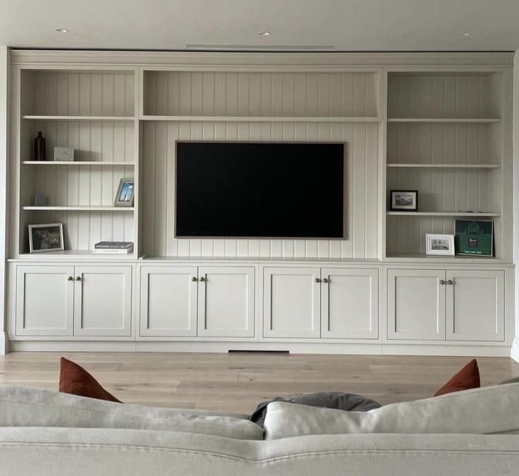 Living room with built-in white shelving unit and a wall-mounted television, surrounded by decorative items such as picture frames and a vase. The foreground features a couch with cushions.