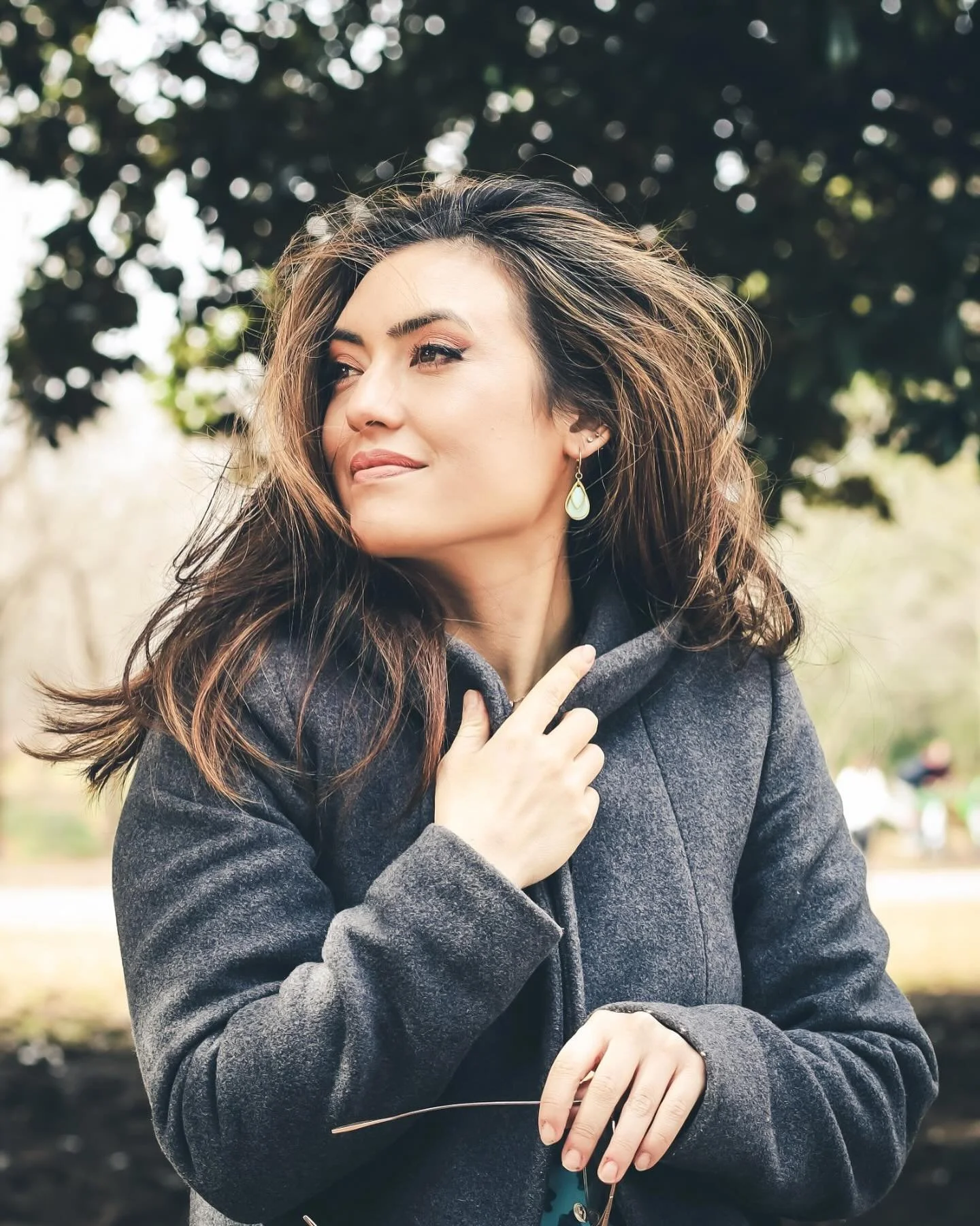 A woman with long brown hair and earrings, wearing a gray coat, standing outdoors with trees in the background, looking to the side with a slight smile.