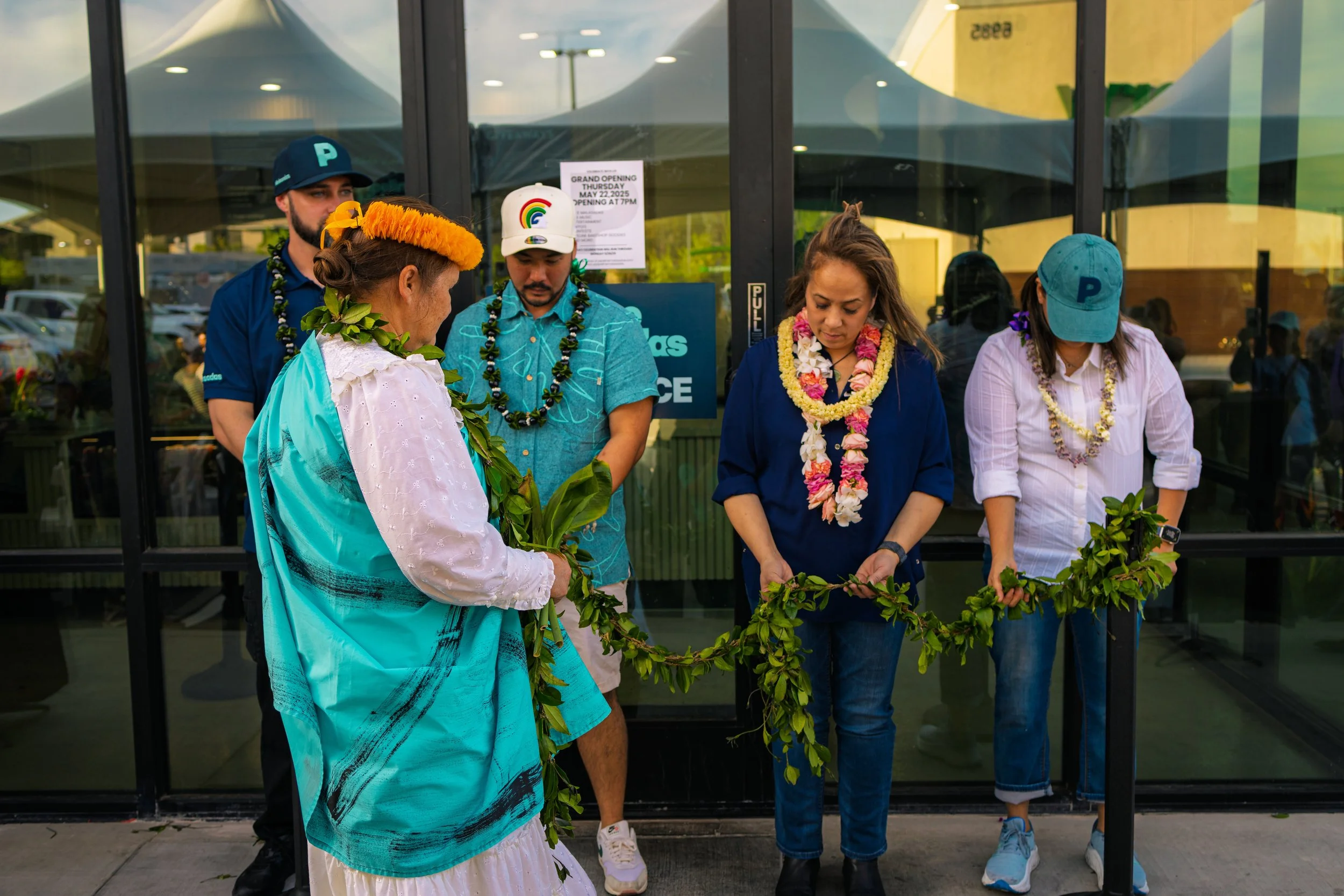 A group of five people participating in a traditional Hawaiian ceremony outside a building. They are wearing leis and holding a floral garland together, with some dressed in casual clothes and one woman in a traditional Hawaiian dress.