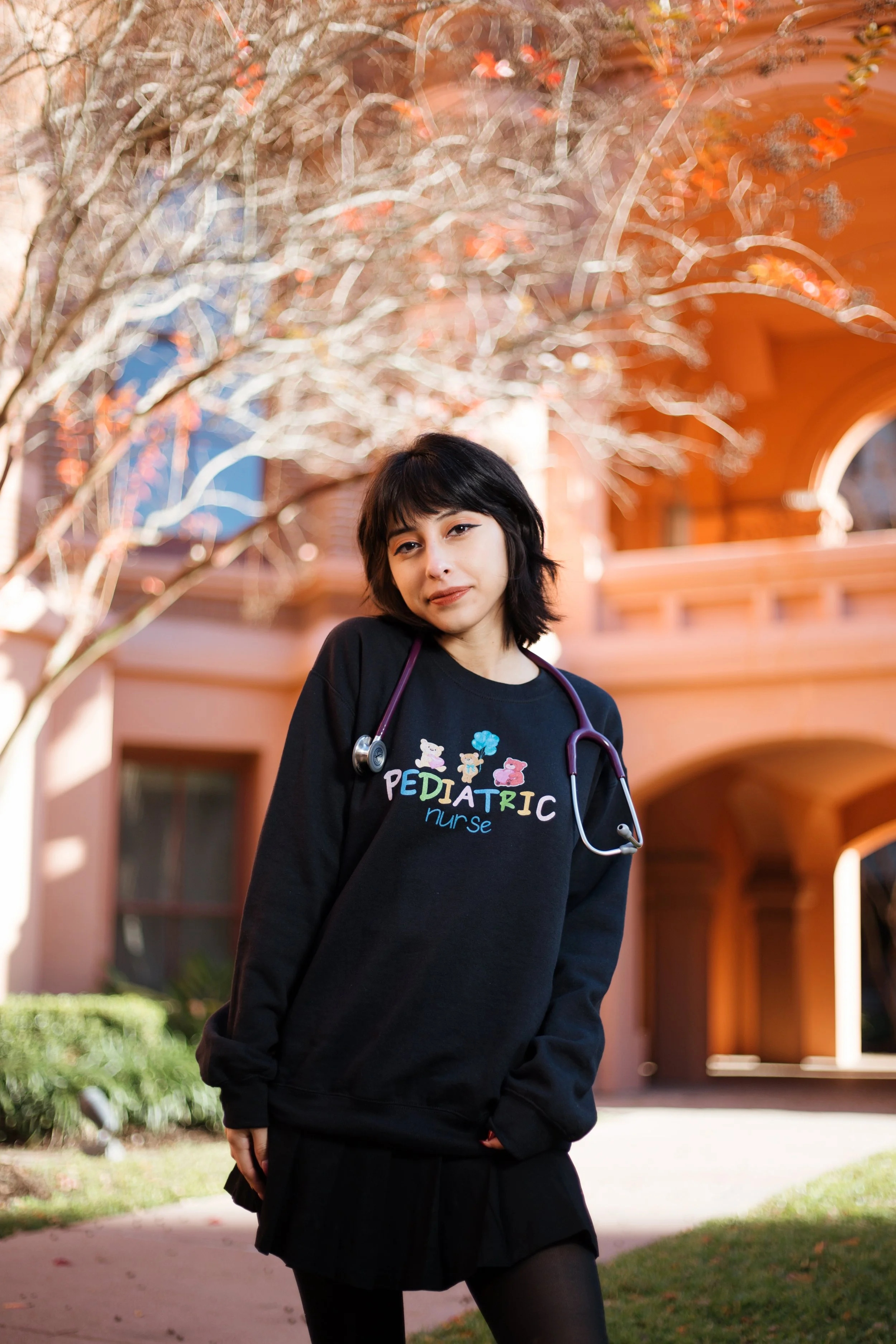 A young woman with dark hair wearing a black sweatshirt that says "PEDIATRIC nurse" sits outside on a sunny day with a pink building and a leafless tree in the background, and a stethoscope around her neck.
