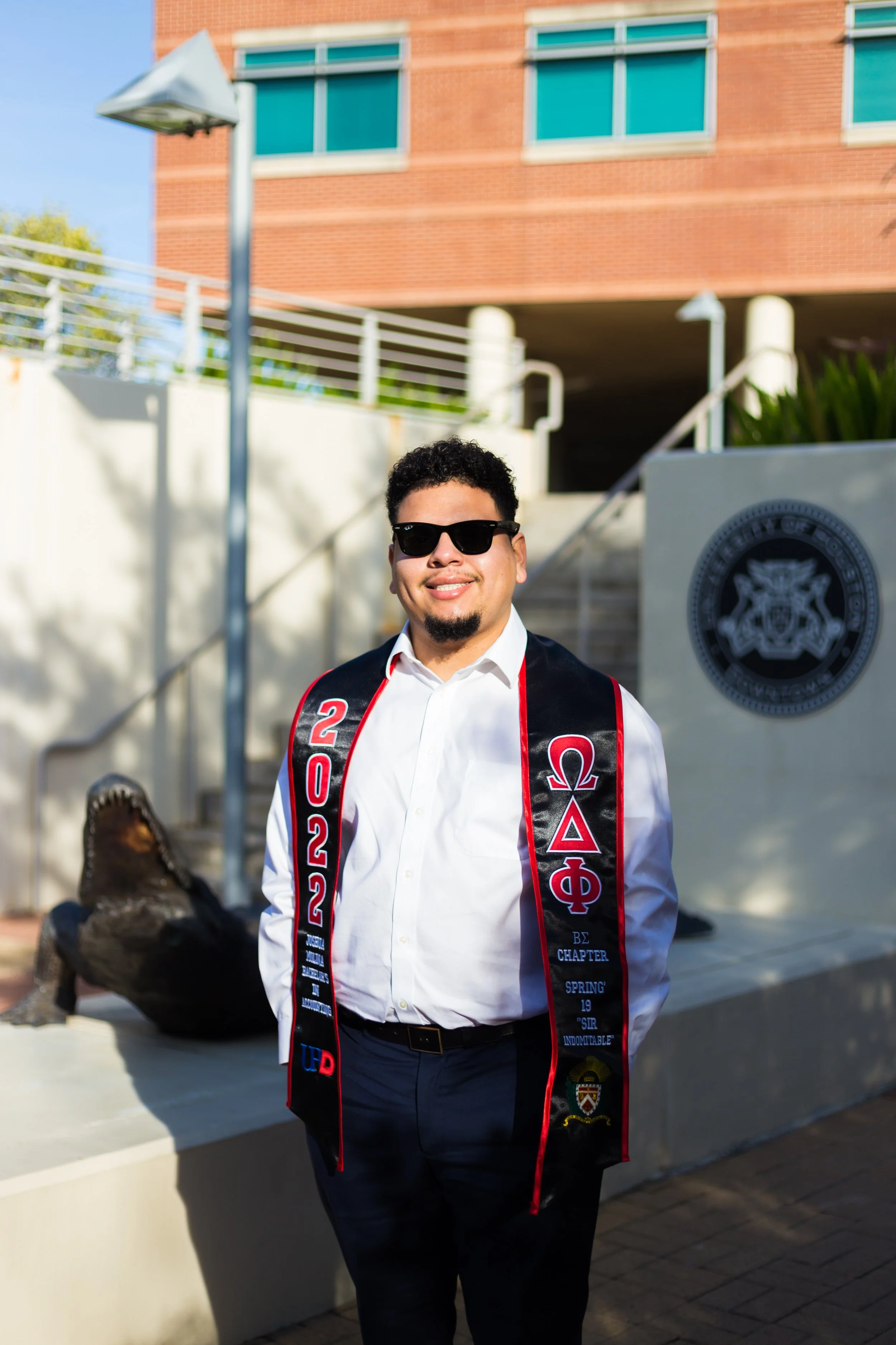 A young man wearing sunglasses, a white shirt, and a graduation sash with fraternity letters and the year 2022 standing outside in front of a building and a sculpture of a crocodile.