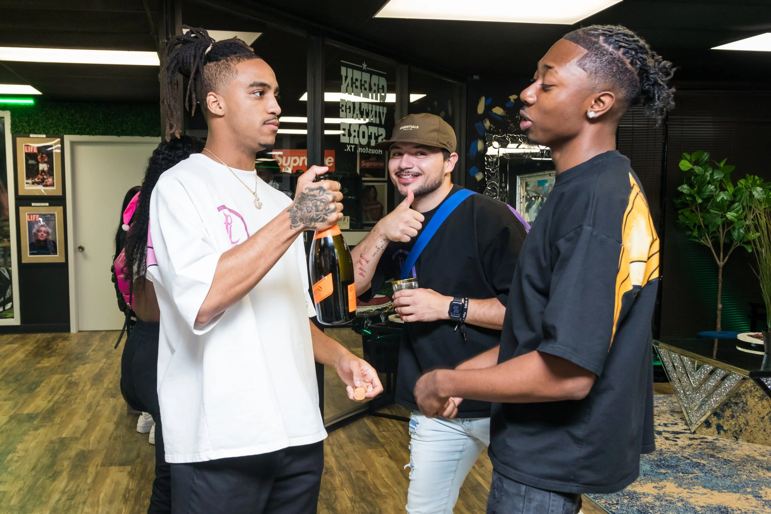 Three young men engaged in conversation at a social gathering, with one holding a bottle of champagne and another holding a drink, inside a modern, decorated room.