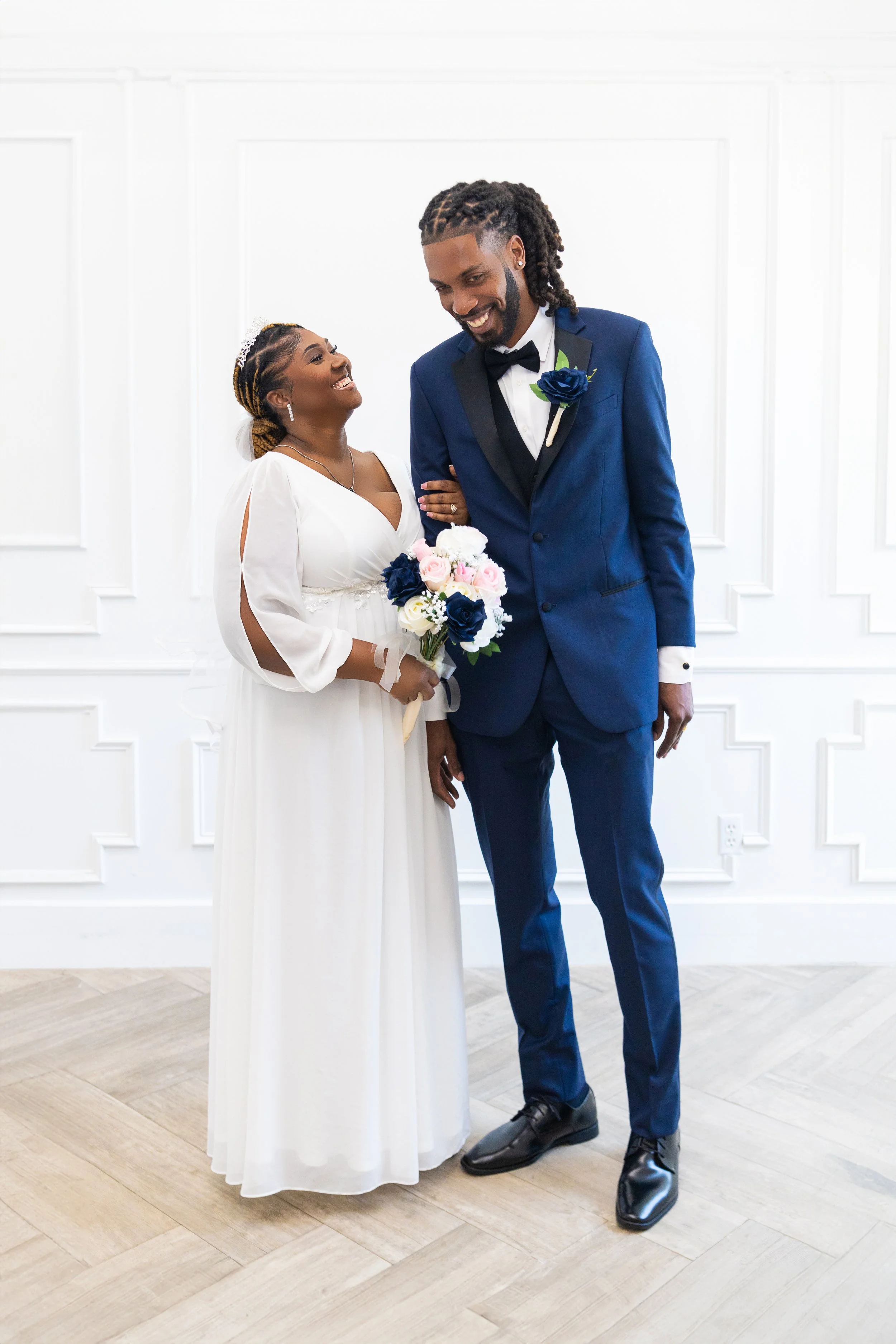 A bride and groom standing side by side on their wedding day, smiling happily, with the bride holding a bouquet of flowers and the groom wearing a dark blue suit with a black bow tie.