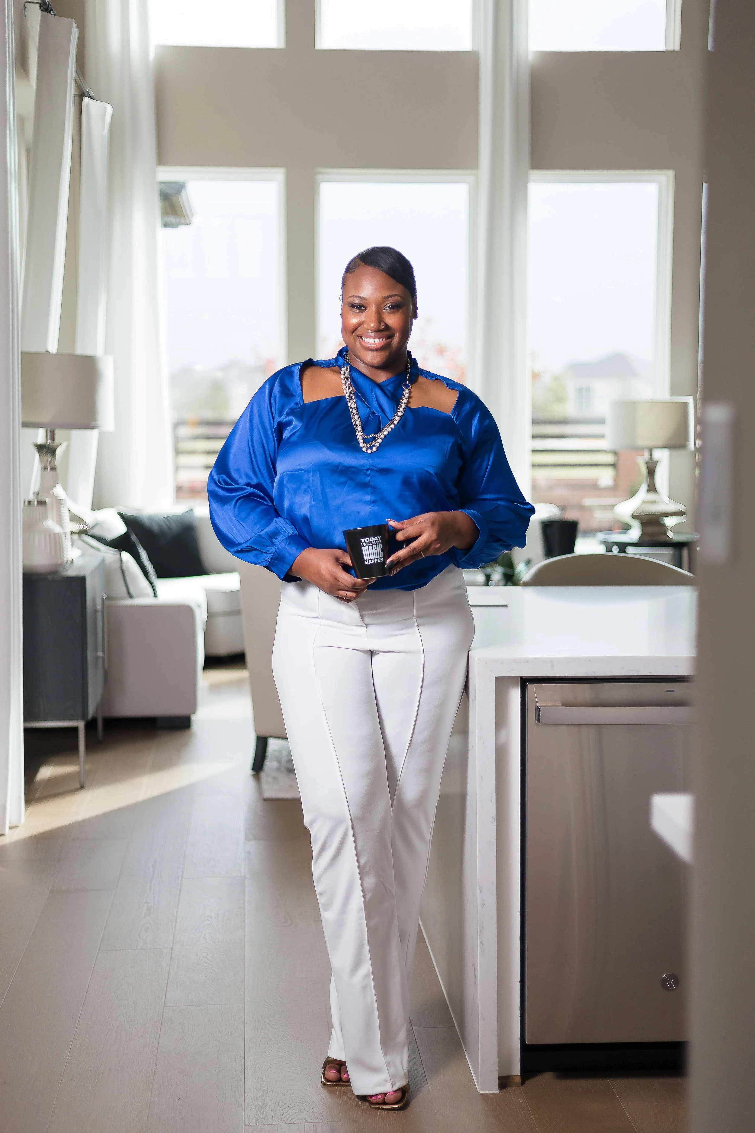 Realtor branding portrait in Katy, Texas by Lev’s Photography featuring a woman in a bright blue blouse and white pants holding a black mug in a modern kitchen with white furniture and large windows