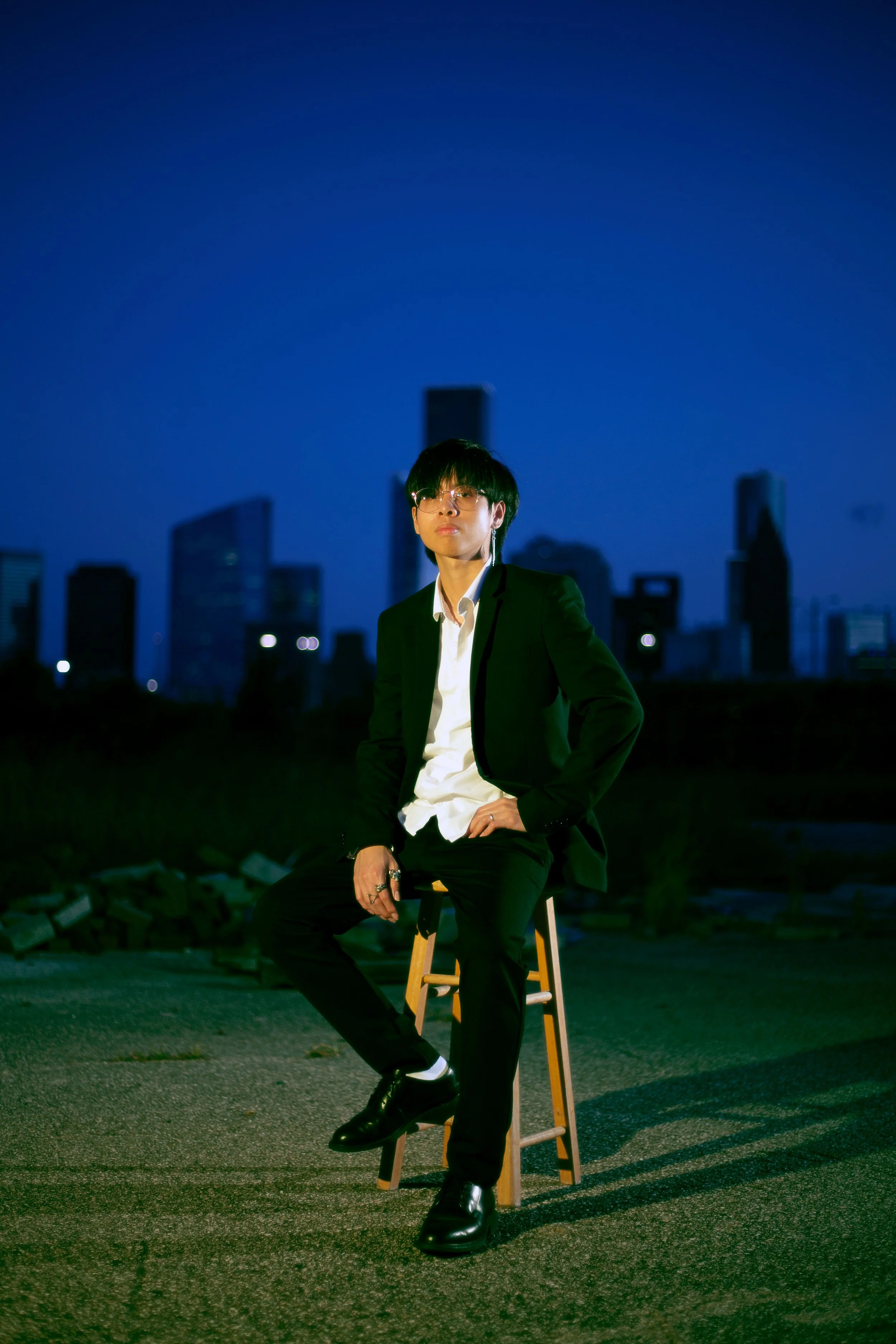 Young man in black suit and white shirt sitting on wooden stool outdoors during nighttime with city skyline in the background.