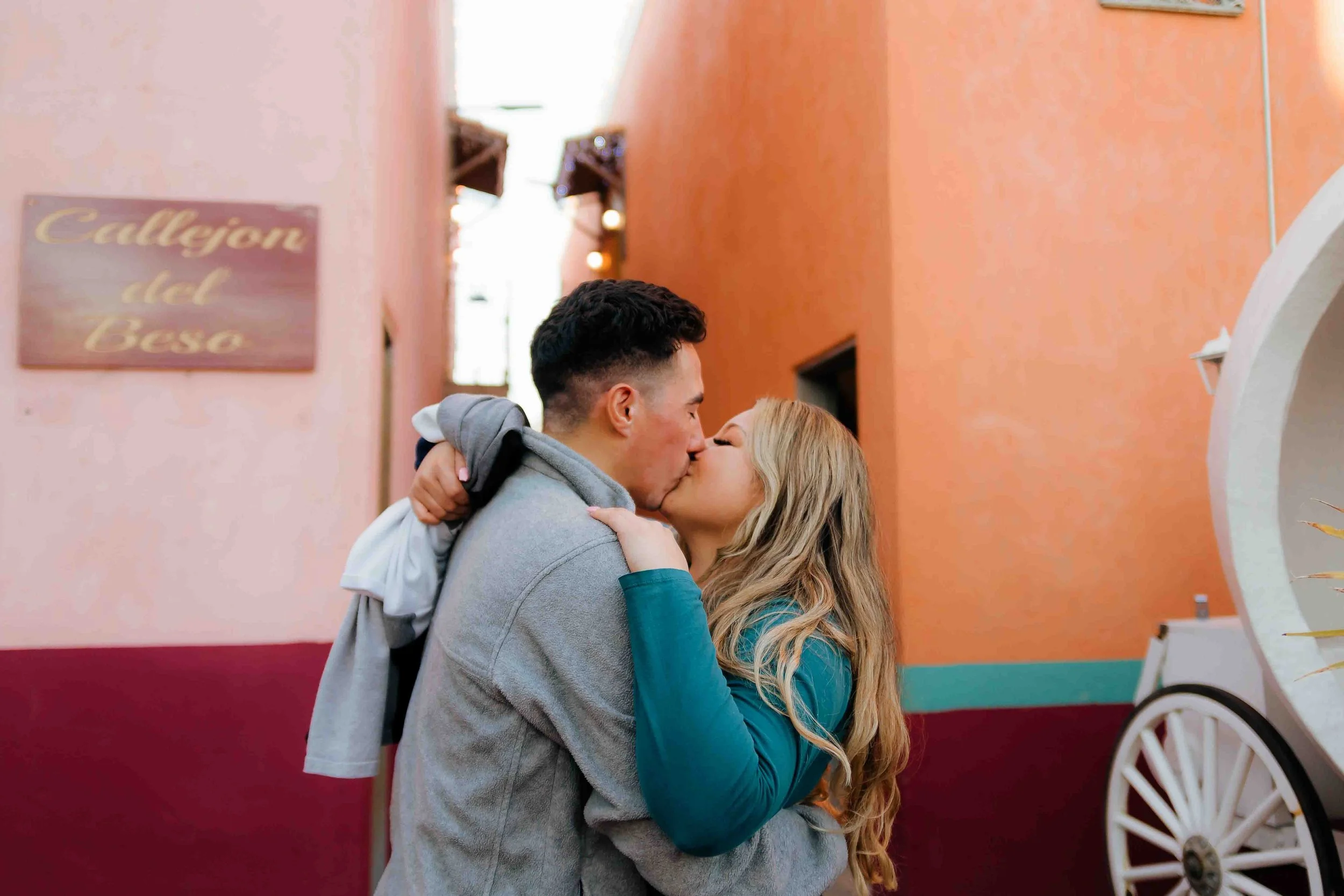 Romantic couple portrait by Lev’s Photography featuring a young couple kissing in a colorful alleyway with pink and orange walls and a “Callejón del Beso” sign in the background