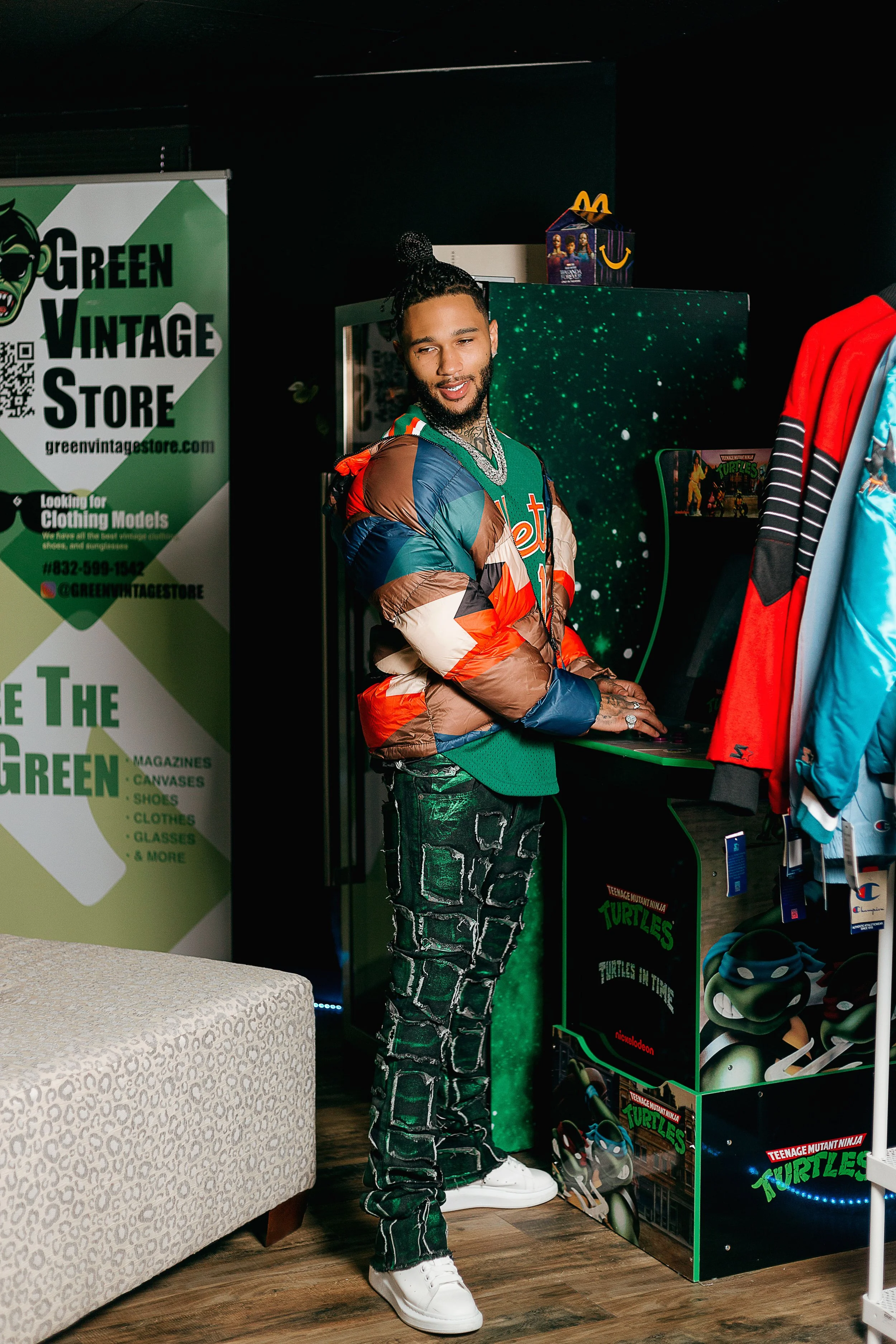 Man with braided hair and tattoos wearing a multicolored puffer jacket and patterned pants standing inside a vintage store, surrounded by Teenage Mutant Ninja Turtles merchandise and clothing racks.