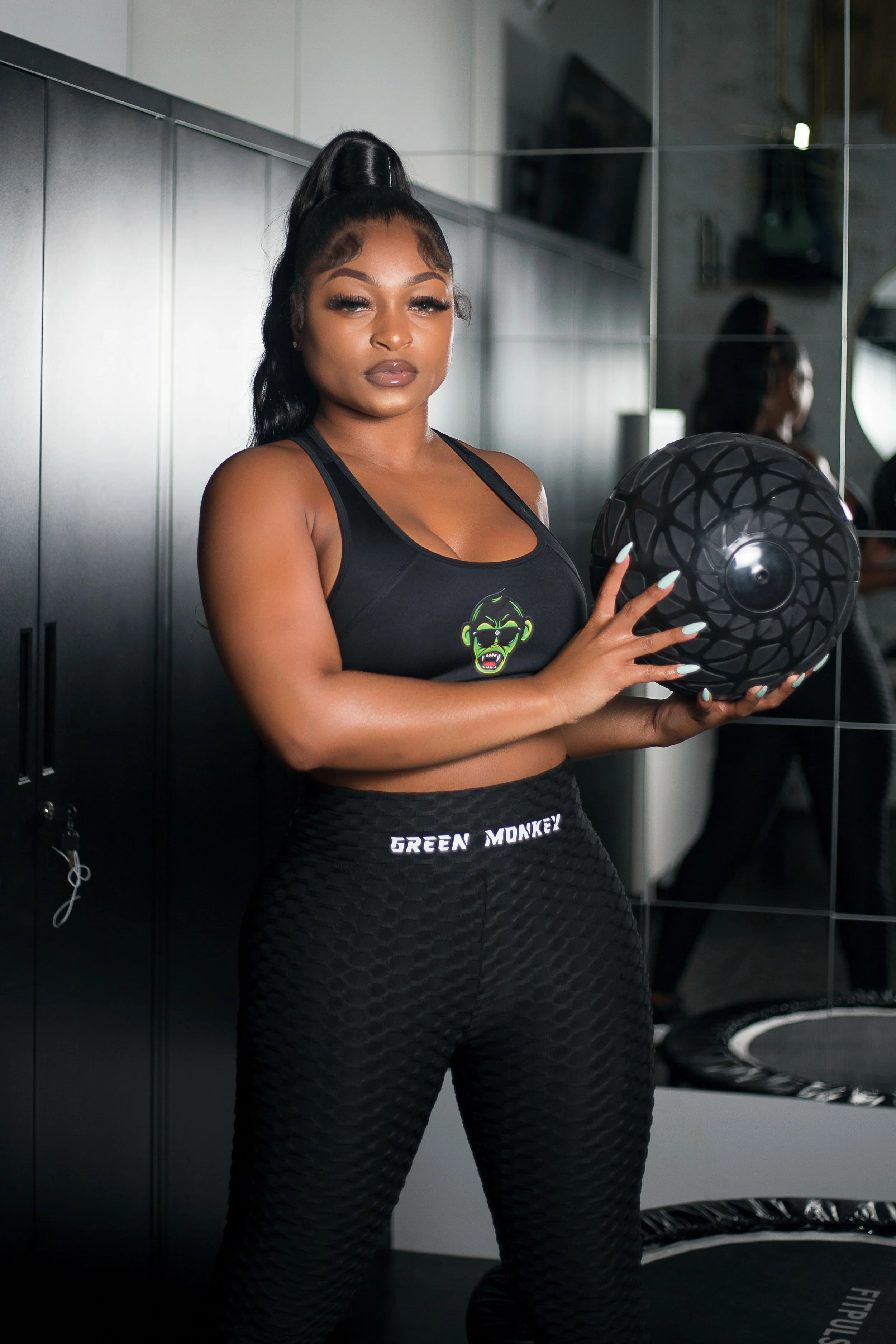 A young woman with long black hair tied in a high ponytail wearing black athletic wear with a green monkey logo, holding a black exercise ball, standing in a gym.