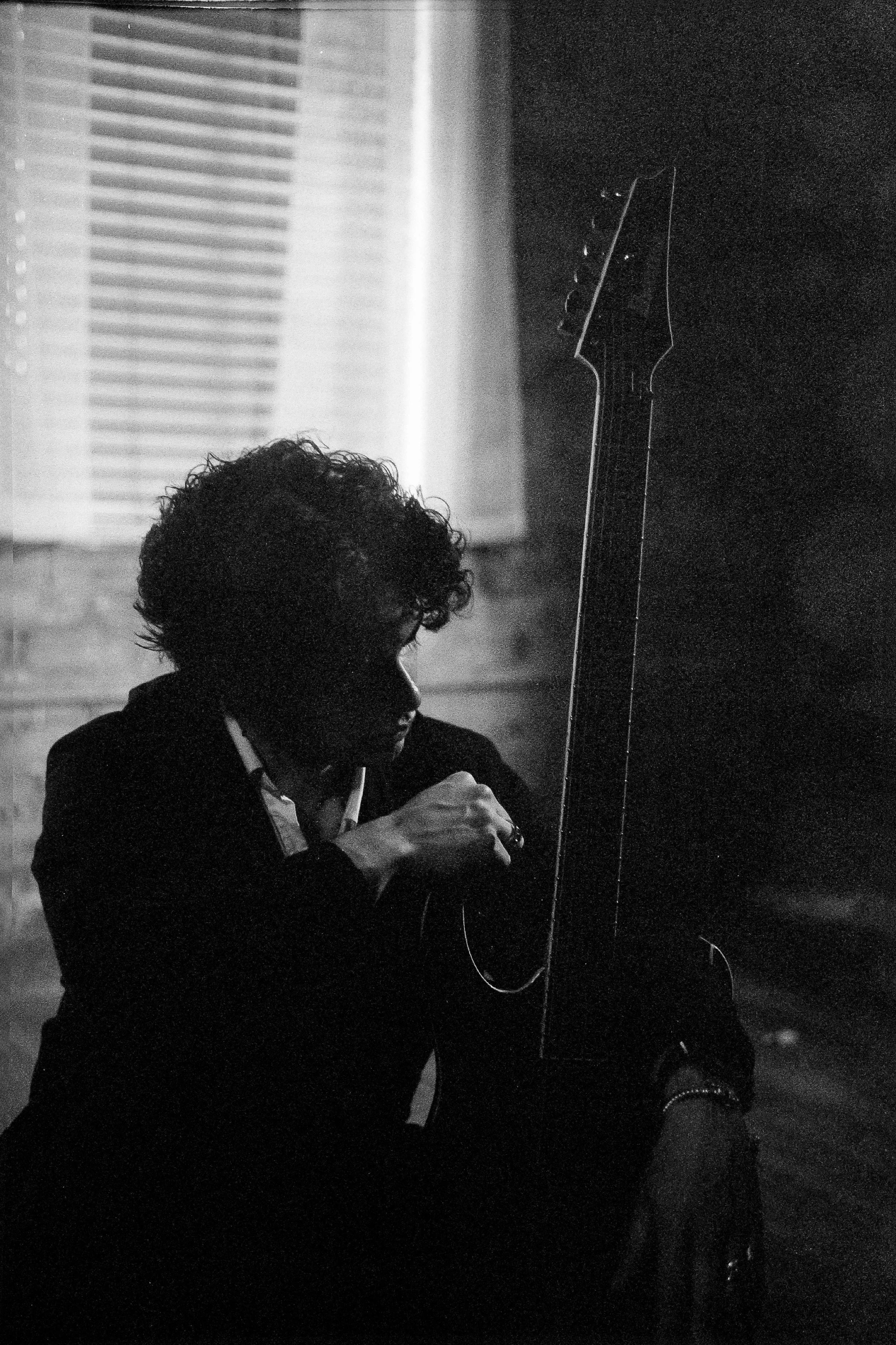 Black and white photo of a person with curly hair sitting in a dark room, holding a guitar and looking down. Light is coming from a window with blinds in the background.