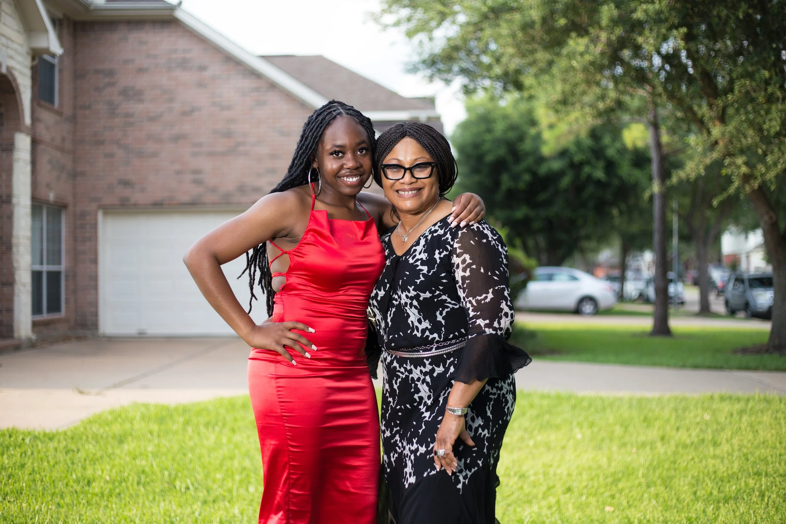 Two women standing on a grass lawn in front of a brick house, smiling and embracing. One woman is wearing a bright red dress with cut-out sides, and the other woman is wearing a black and white patterned dress with glasses.