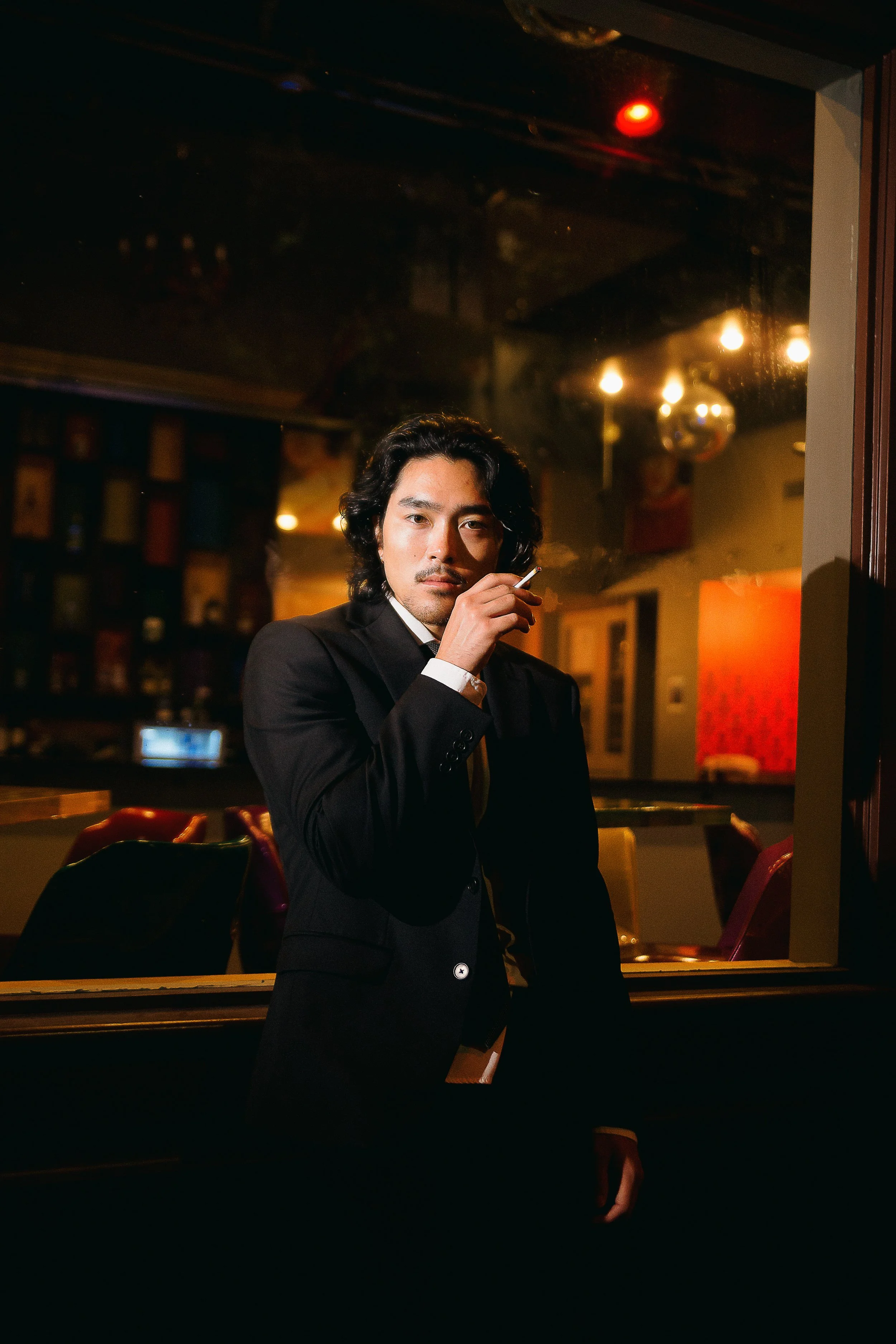 Man in black suit holding a cigarette in a dimly lit indoor setting, looking directly at the camera.