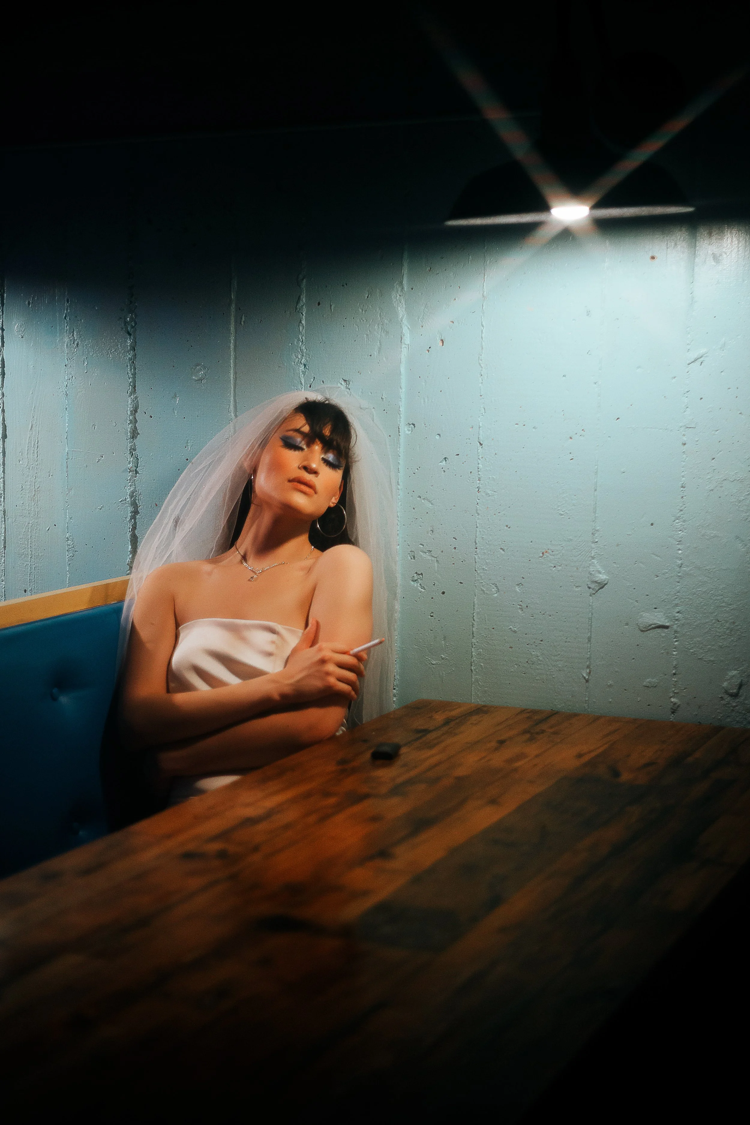 A woman dressed as a bride with a veil and strapless dress sitting alone at a wooden table in a dimly lit room, holding a cigarette.