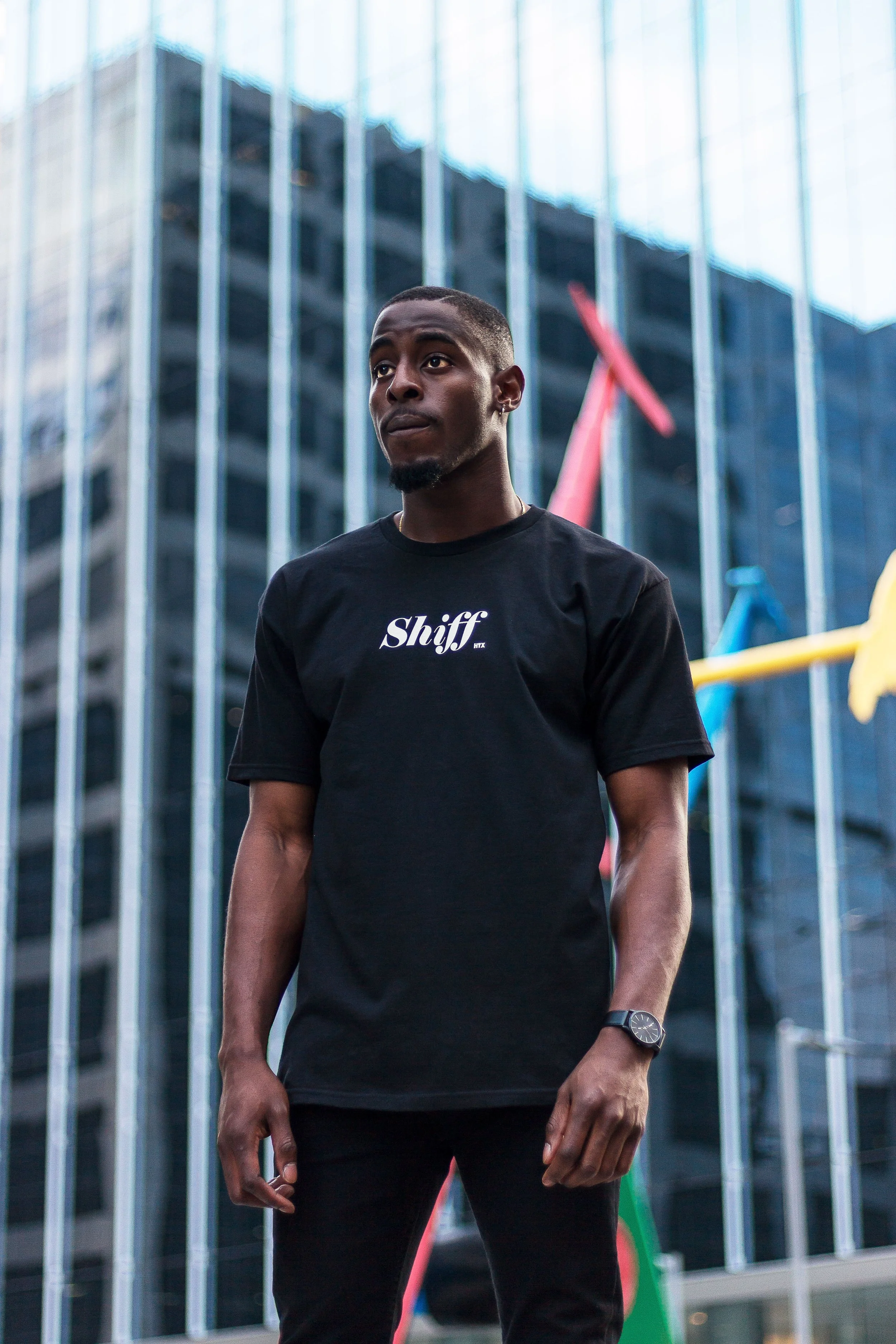 A young man standing outdoors in front of a modern glass building, wearing a black t-shirt with the text 'Shiff' on it, and a wristwatch, with colorful abstract sculptures in the background.