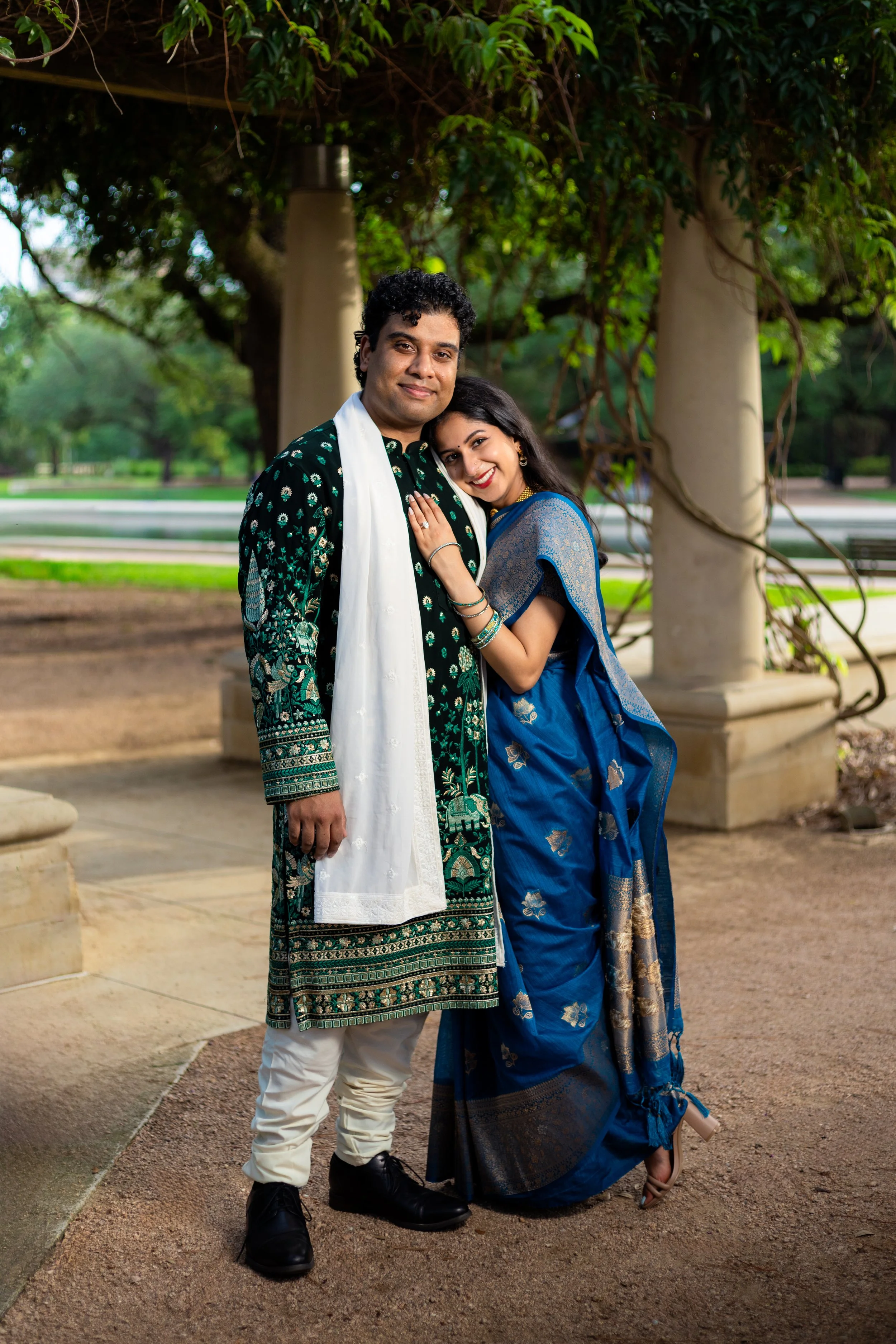 A smiling couple in traditional Indian attire standing outdoors under a tree with columns in the background. The woman is touching the man's chest and wearing a blue saree with gold accents, while the man is dressed in a dark green and black kurta wi