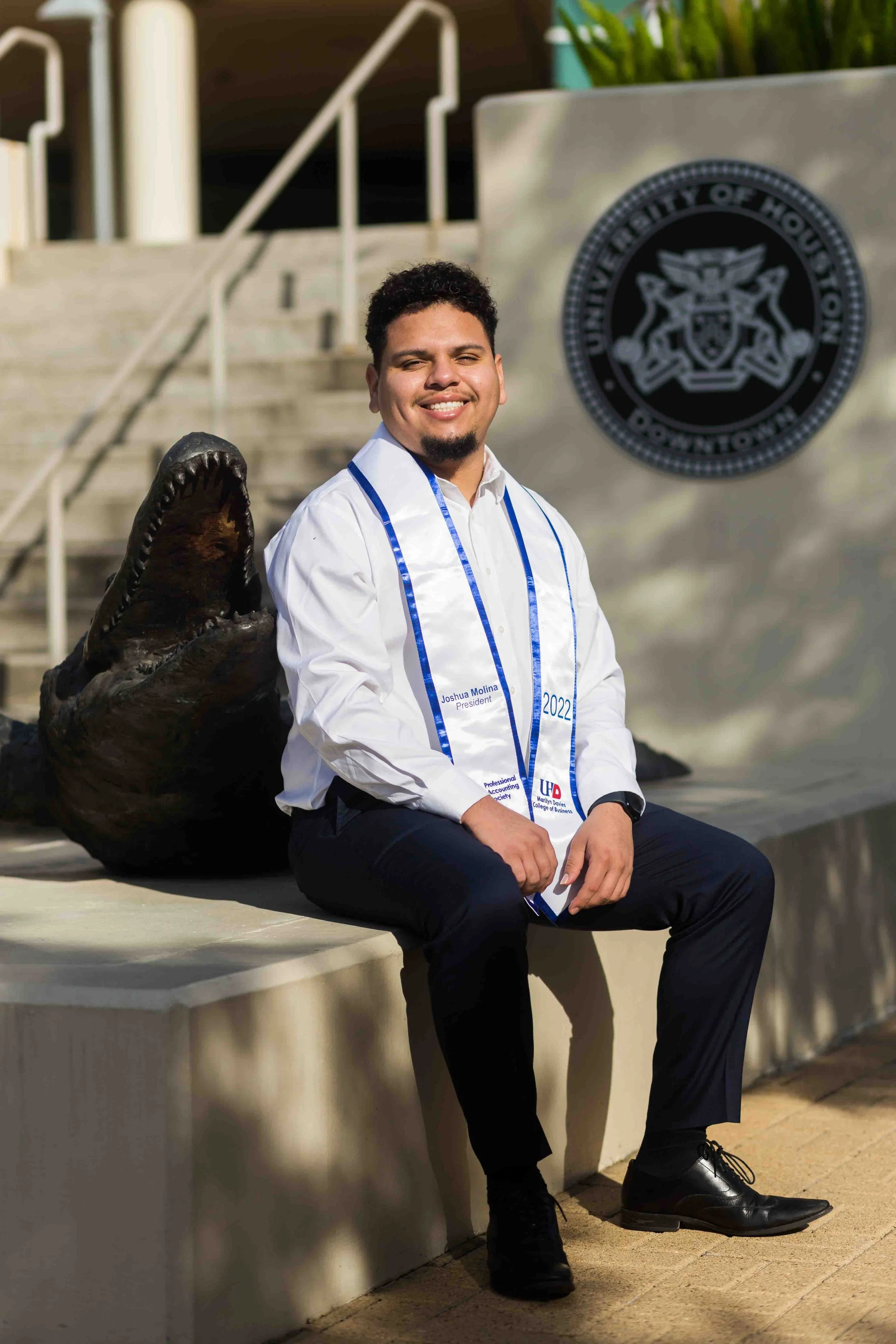 Graduation portrait of a young man from the Marilyn Davies College of Business at the University of Houston–Downtown, wearing formal attire with a diploma ribbon, sitting on a bench outdoors, smiling, with an alligator statue and the UHD seal in the 