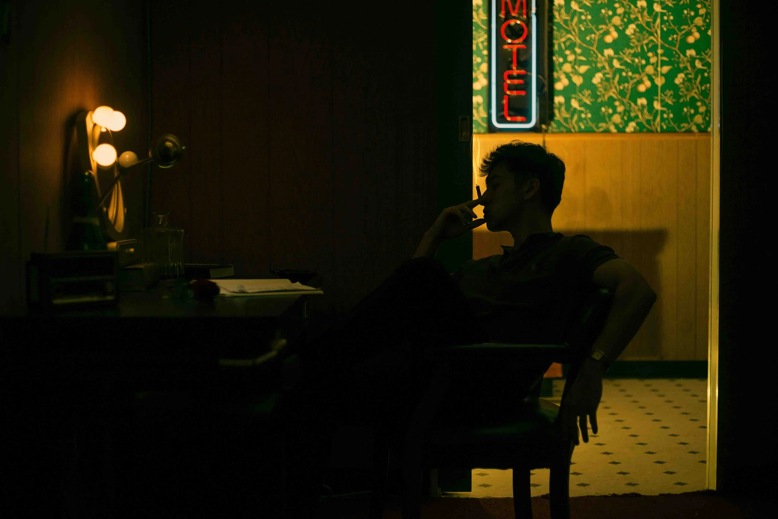 Silhouette of a young man smoking inside a dim motel room with a glowing neon OPEN sign