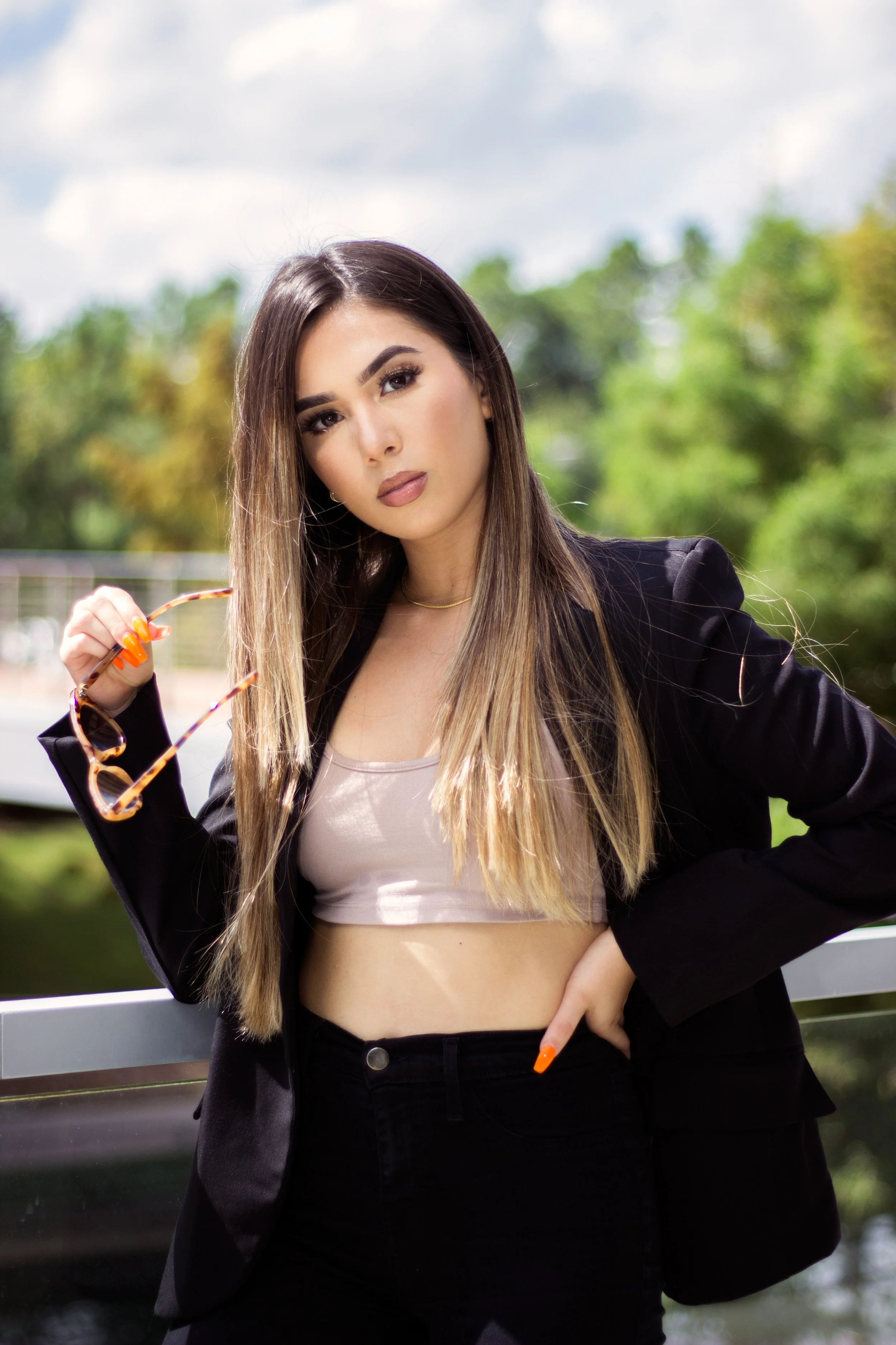 A young woman with long ombre hair, wearing a black blazer, beige crop top, and black pants, holding sunglasses outdoors with a blurred background of trees and a cloudy sky.