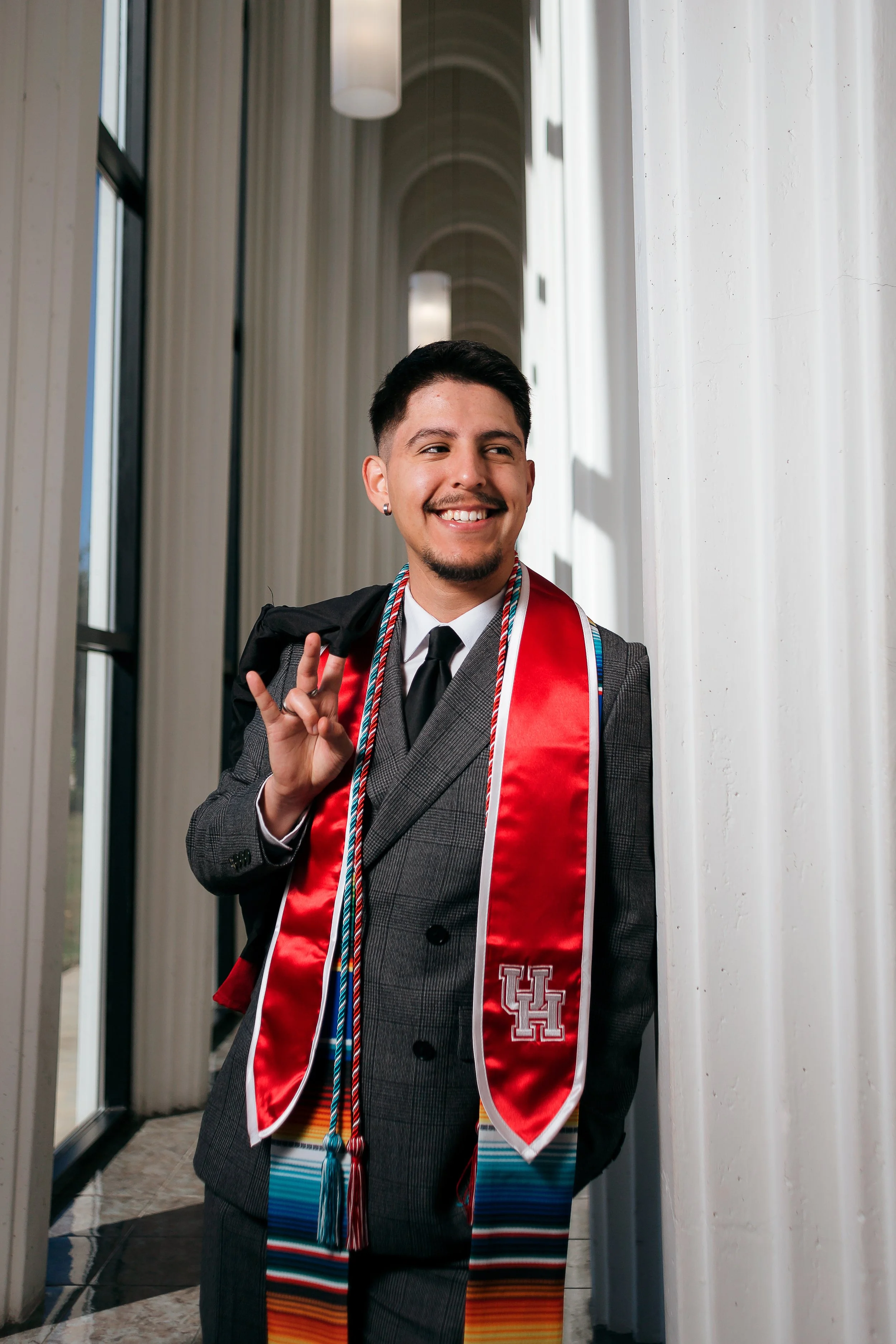 Smiling man in a suit with a black backpack, graduation stole, and honor cords, standing in a modern corridor.
