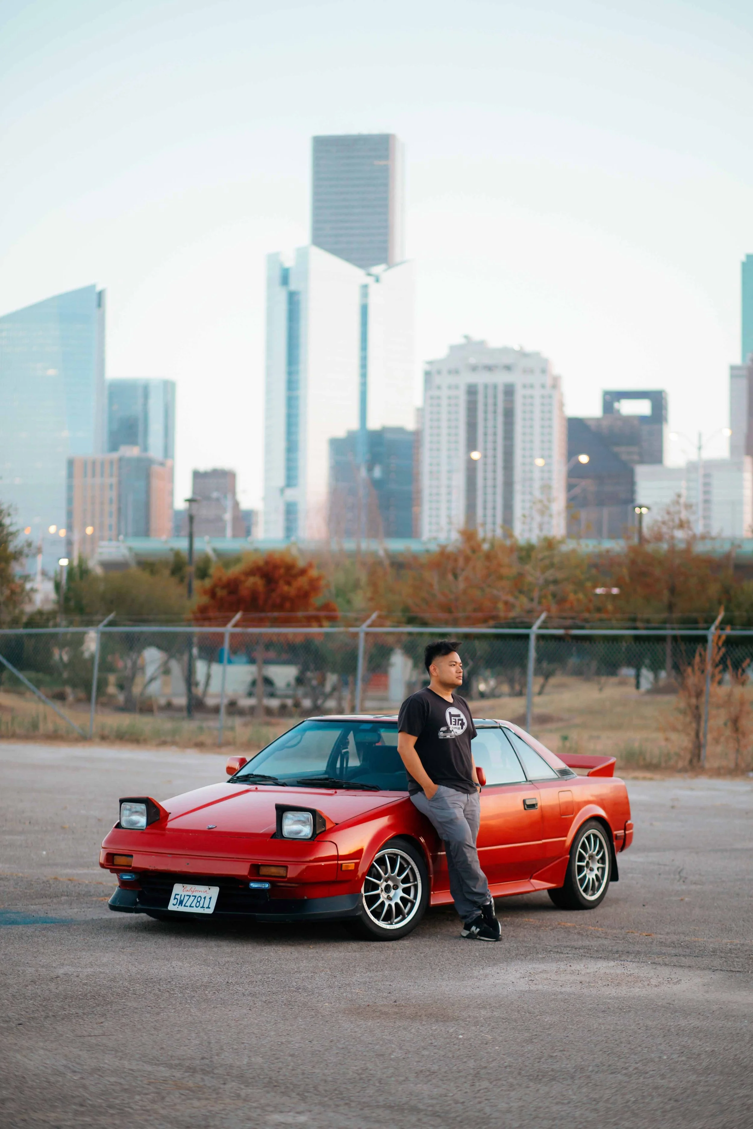 Houston skyline portrait session by Lev’s Photography featuring a man standing beside his red Toyota MR2 in an open parking lot with downtown skyscrapers in the background