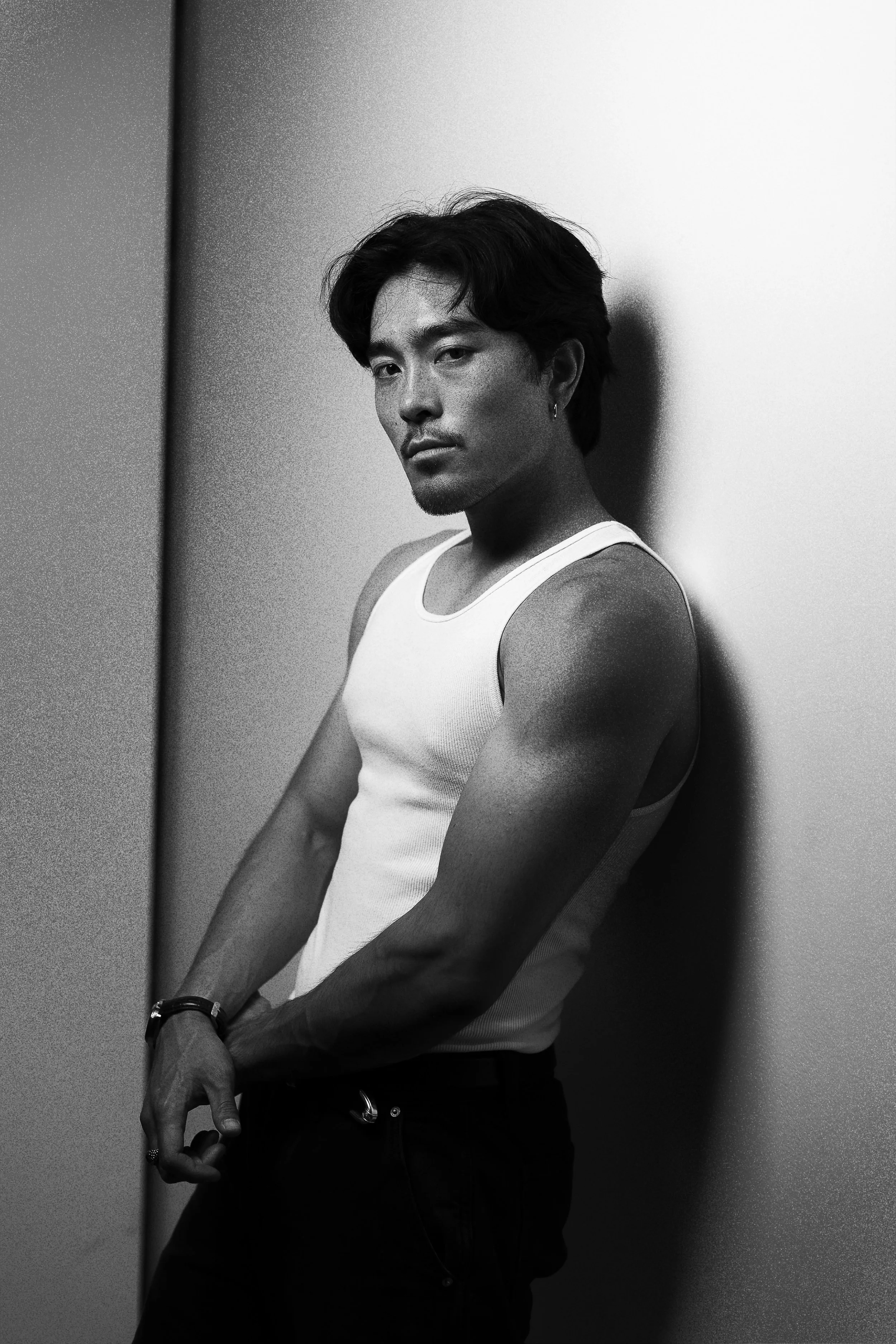 Black and white photo of a man with dark hair wearing a white tank top, standing against a wall with a serious expression.