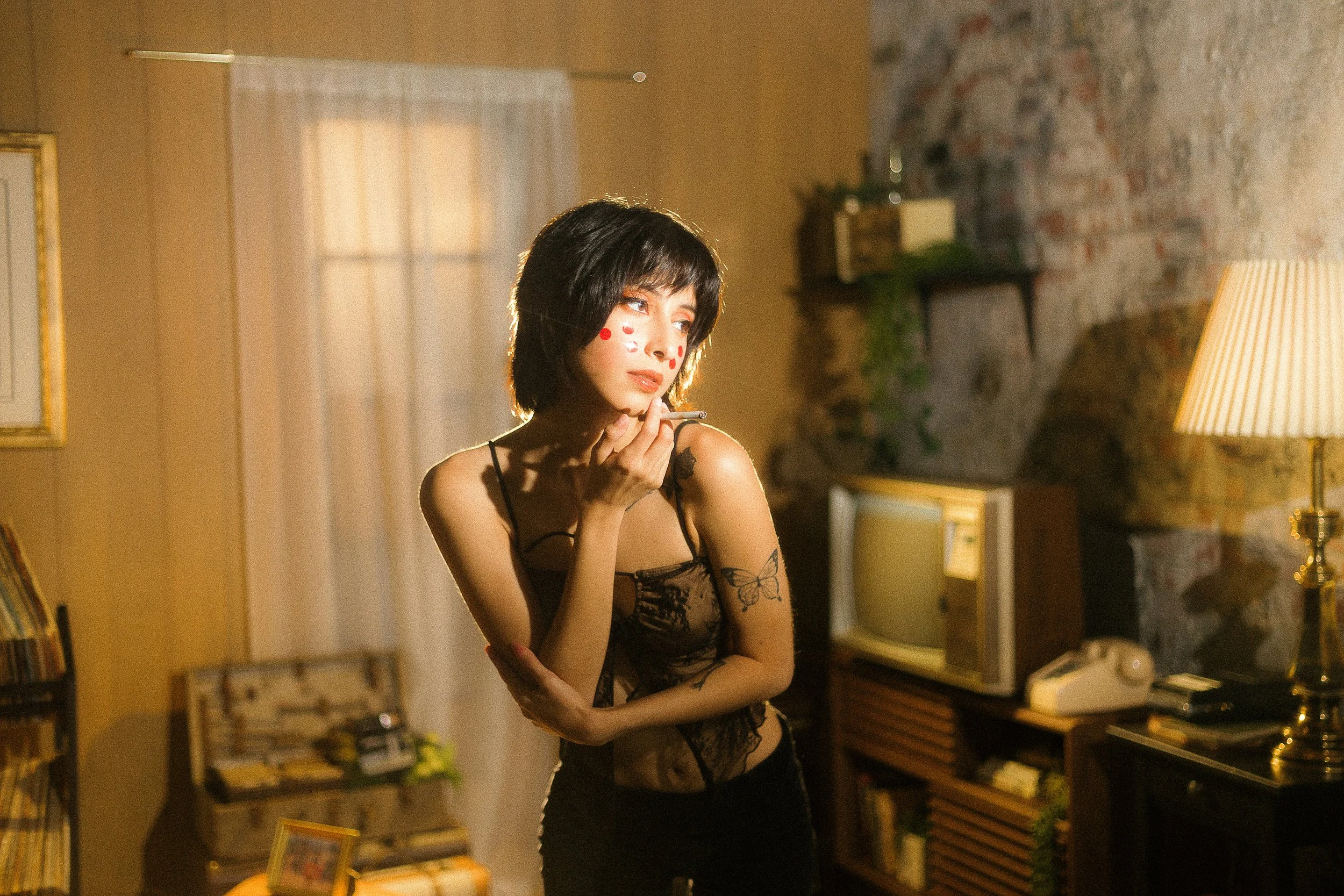 A woman with short dark hair, wearing black lace top and black pants, standing in a dimly lit living room. She has red dots on her face and a butterfly tattoo on her upper arm, holding a cigarette near her lips while looking to the side.
