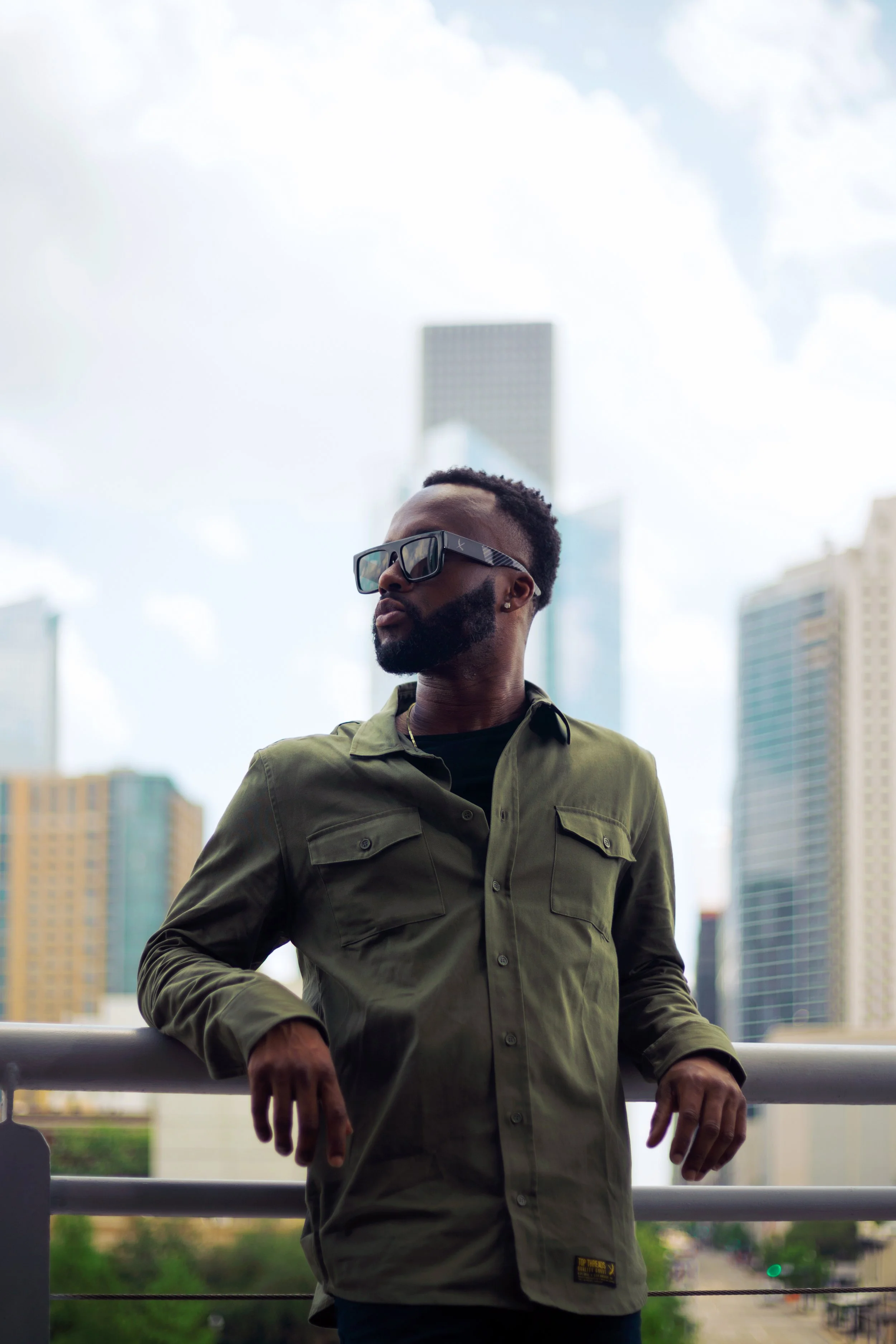 A man with sunglasses and a beard wearing an olive green jacket stands outdoors on a balcony with a city skyline in the background.