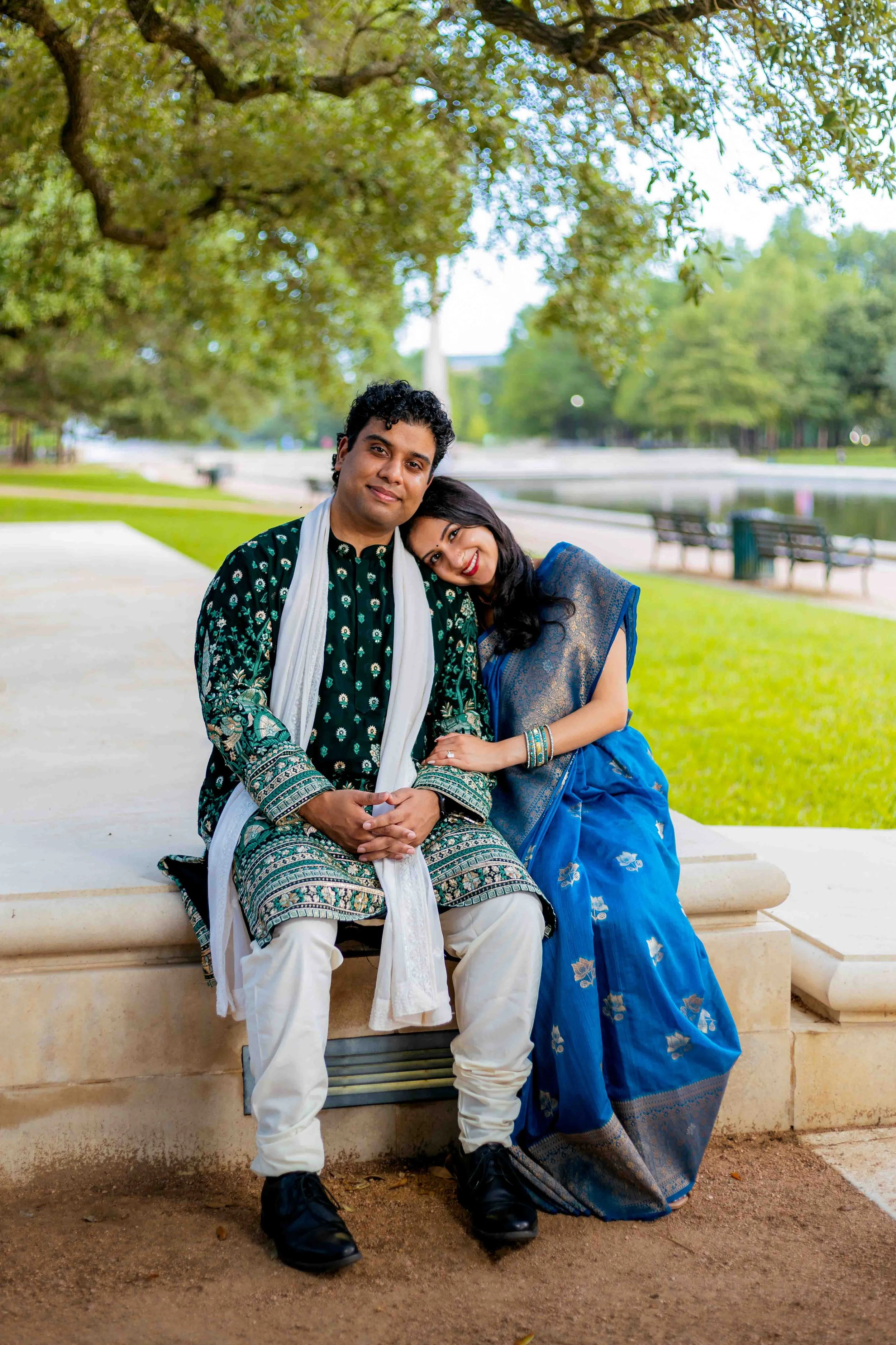 Cultural couple portrait in Houston, Texas by Lev’s Photography featuring a couple dressed in traditional Indian attire sitting on a bench in a park with trees, a fountain, and water in the background