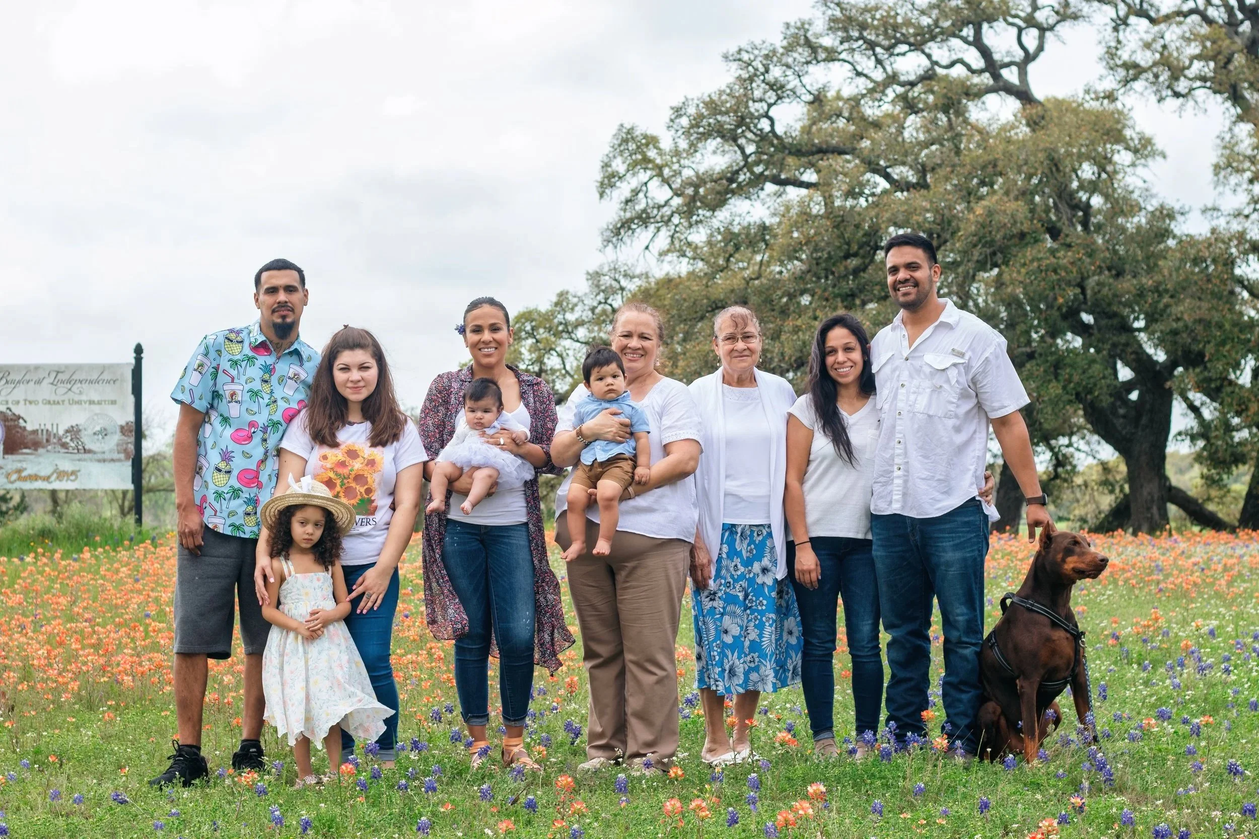 Family photo of ten people and a dog standing in a field of colorful flowers with a tree and a sign in the background.