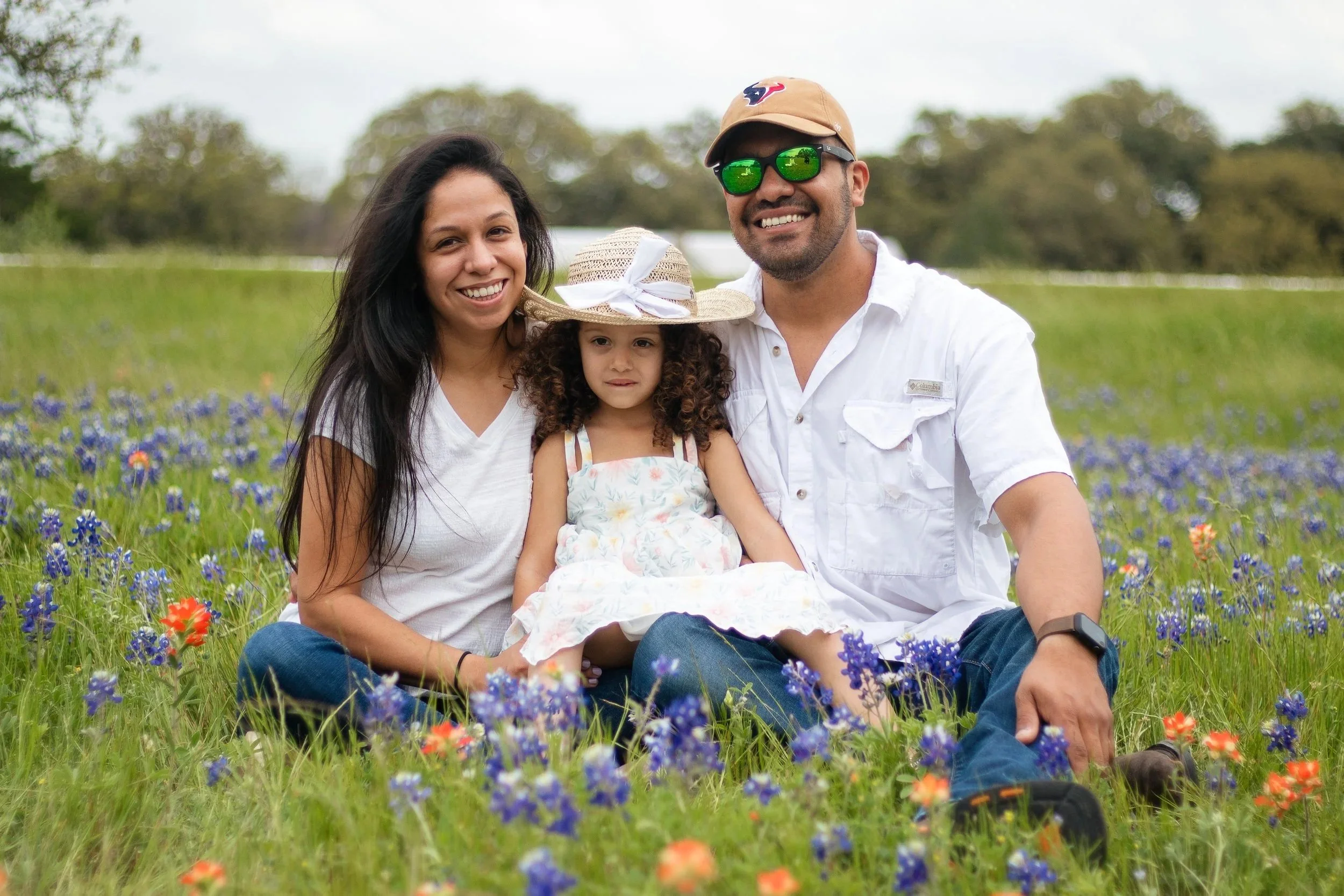 A family of three sitting outdoors in a field of flowers, smiling at the camera. The woman has long dark hair, wearing a white t-shirt. The man has a beard, sunglasses, a beige cap with a logo, and a white shirt. The girl is wearing a straw hat with