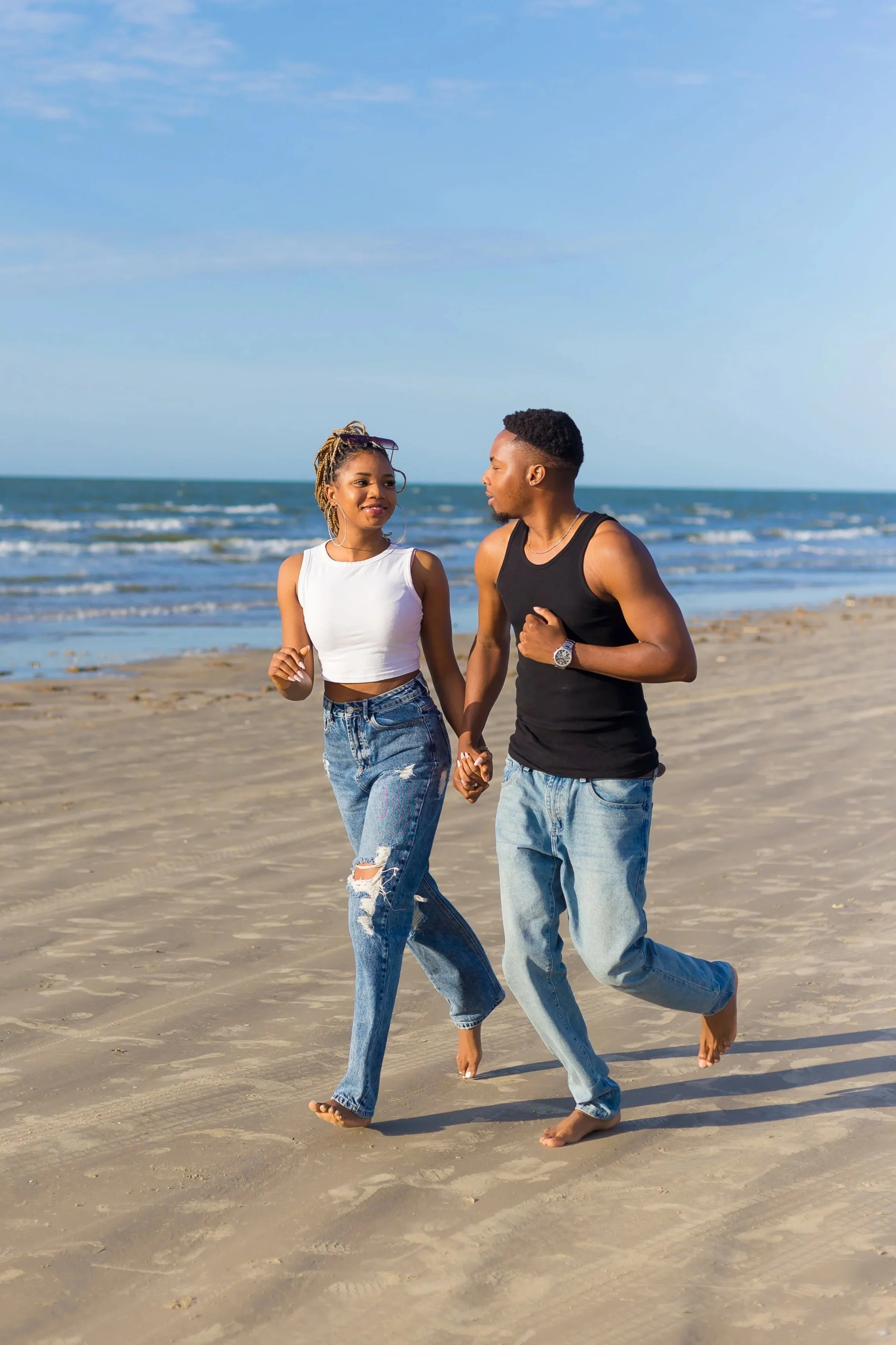 A couple walking hand-in-hand on the beach, with the ocean and blue sky in the background.