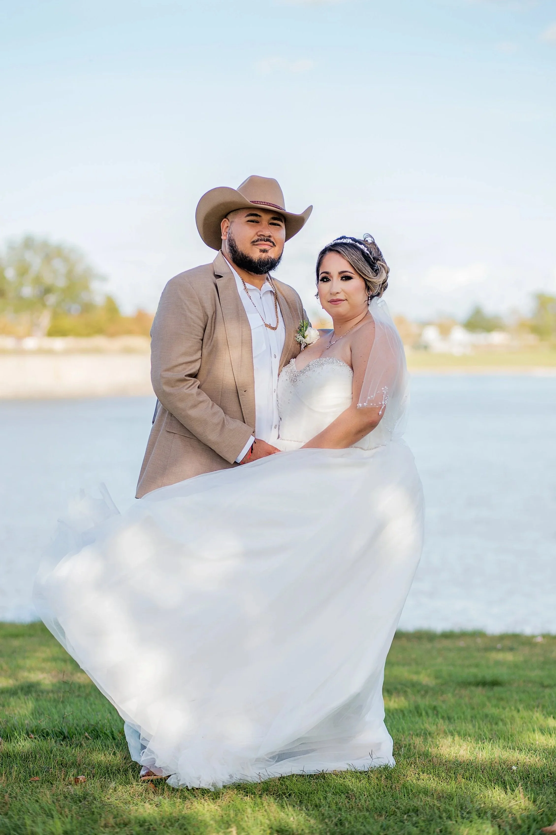 A newlywed couple stands outdoors by the water, with a man wearing a beige suit and cowboy hat, and a woman in a white wedding gown with a veil.