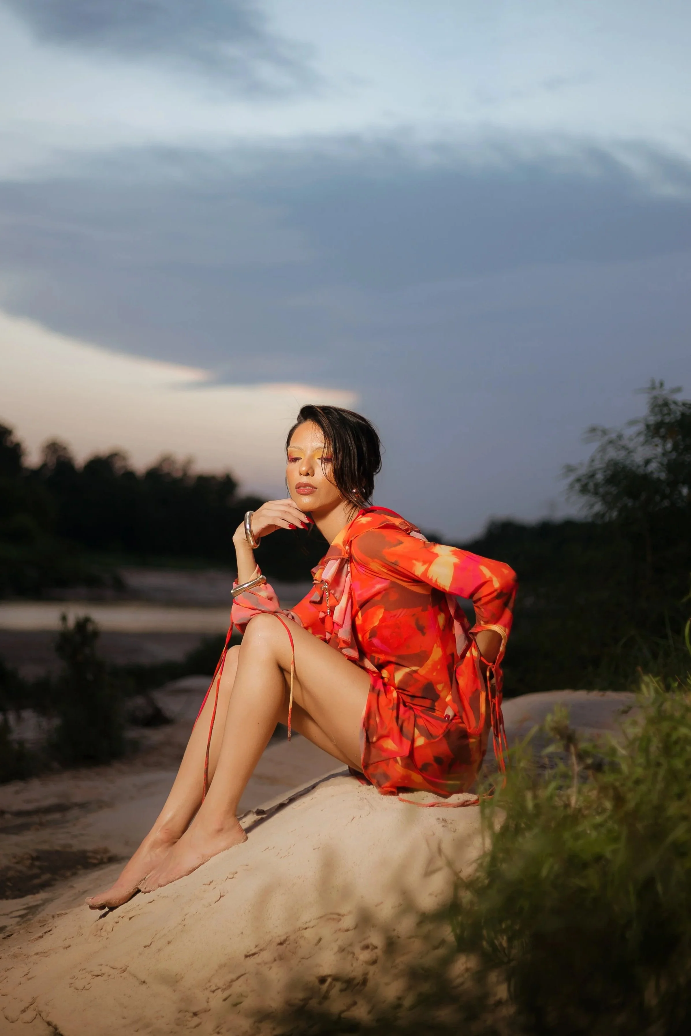 A woman in a vibrant orange and red dress sits on a sandy dune outdoors during dusk with a cloudy sky in the background.