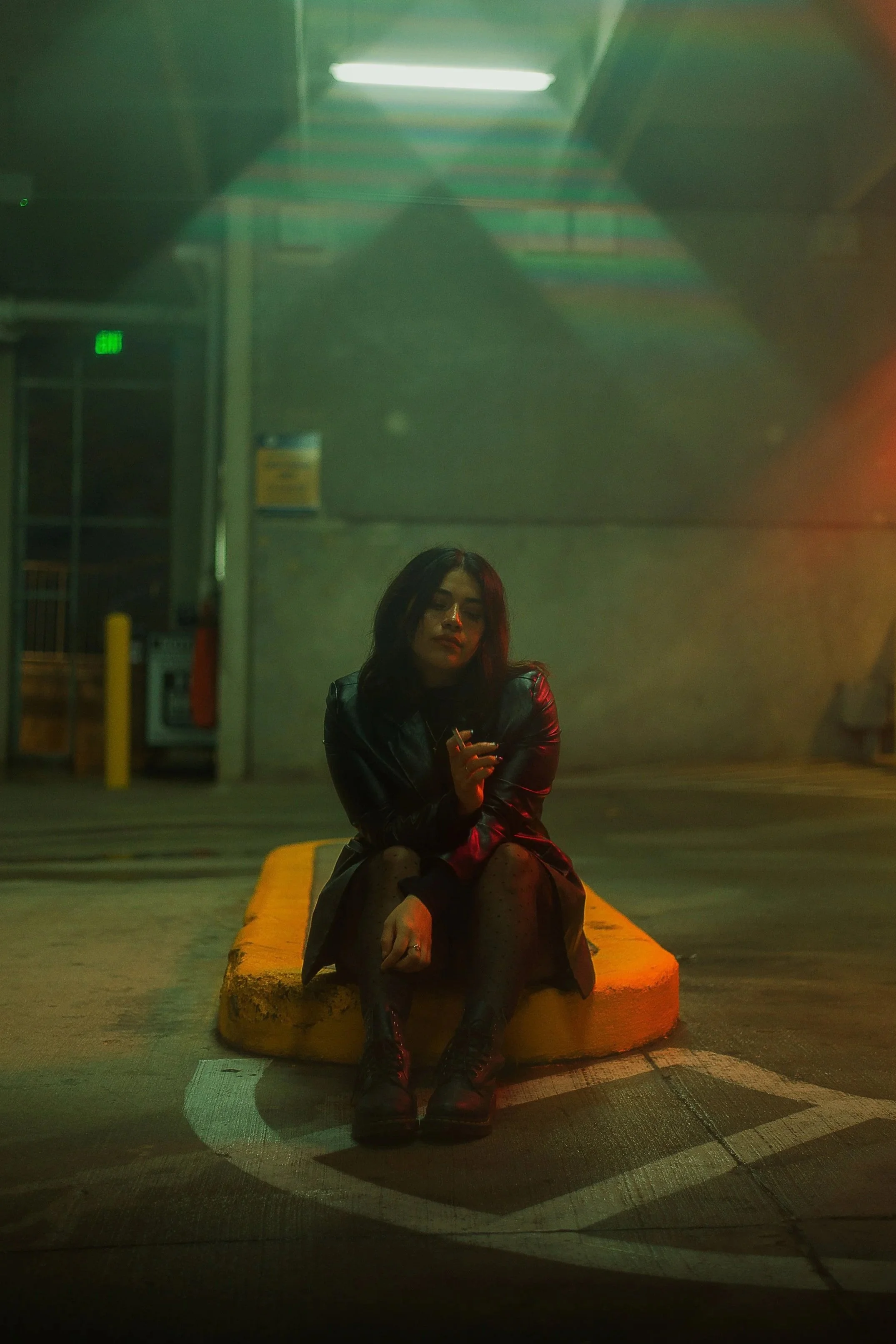 A woman sitting on a yellow curb in a parking lot at night, holding a cigarette, wearing a black leather jacket, black dress, and combat boots, illuminated by multicolored streetlights.
