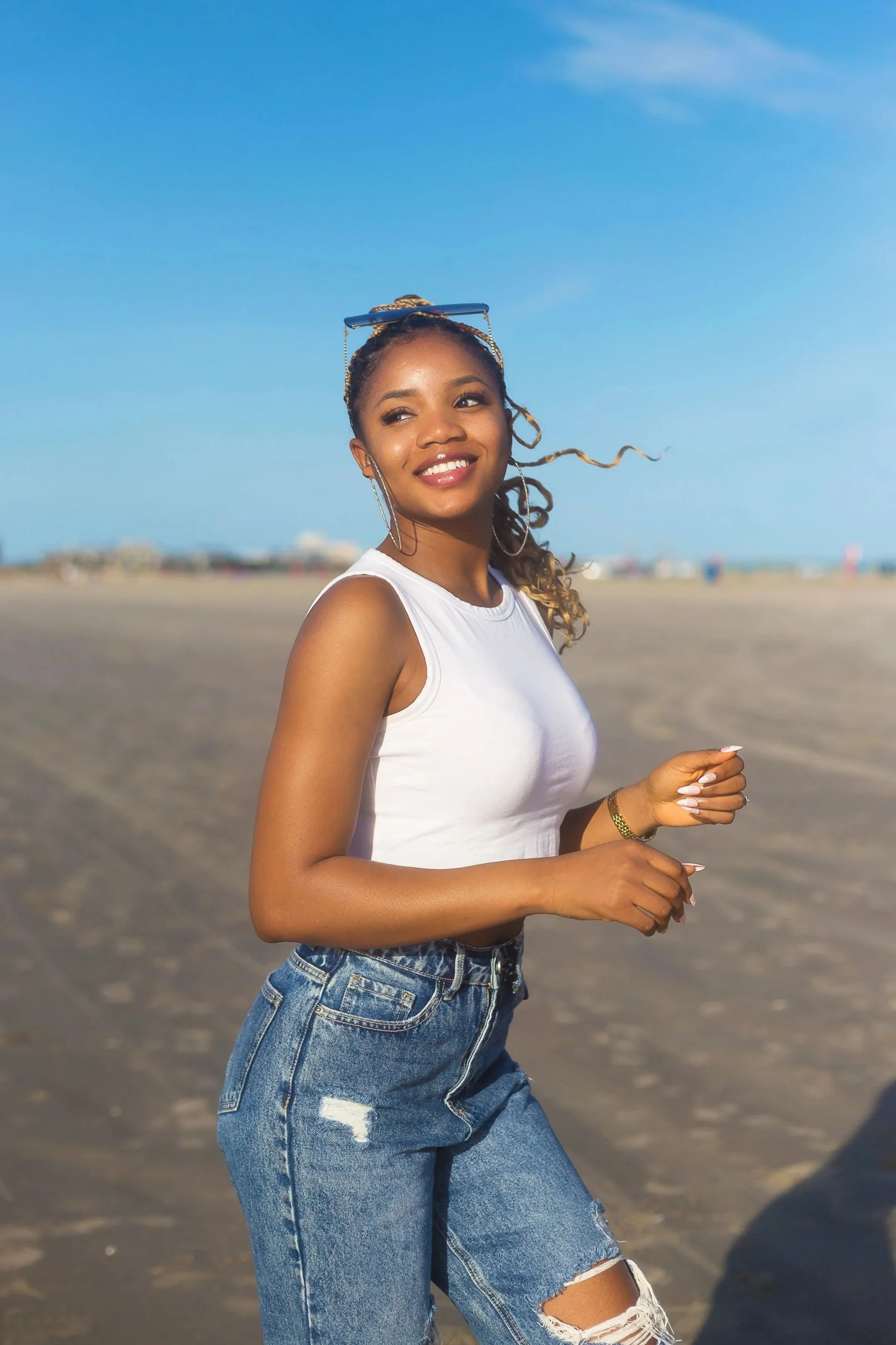 A young woman with long curly hair and sunglasses on her head, smiling while walking on a sandy beach under a clear blue sky.