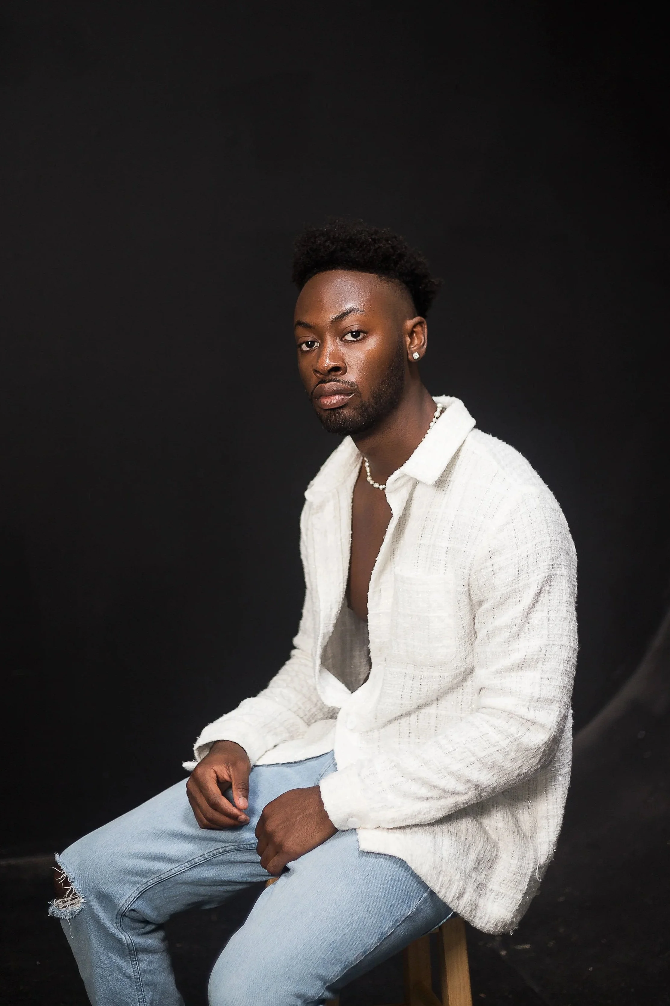 A young man with dark skin and styled hair sitting on a stool against a black background wearing a white open shirt, light blue ripped jeans, and accessorized with earrings, a necklace, and a bracelet.