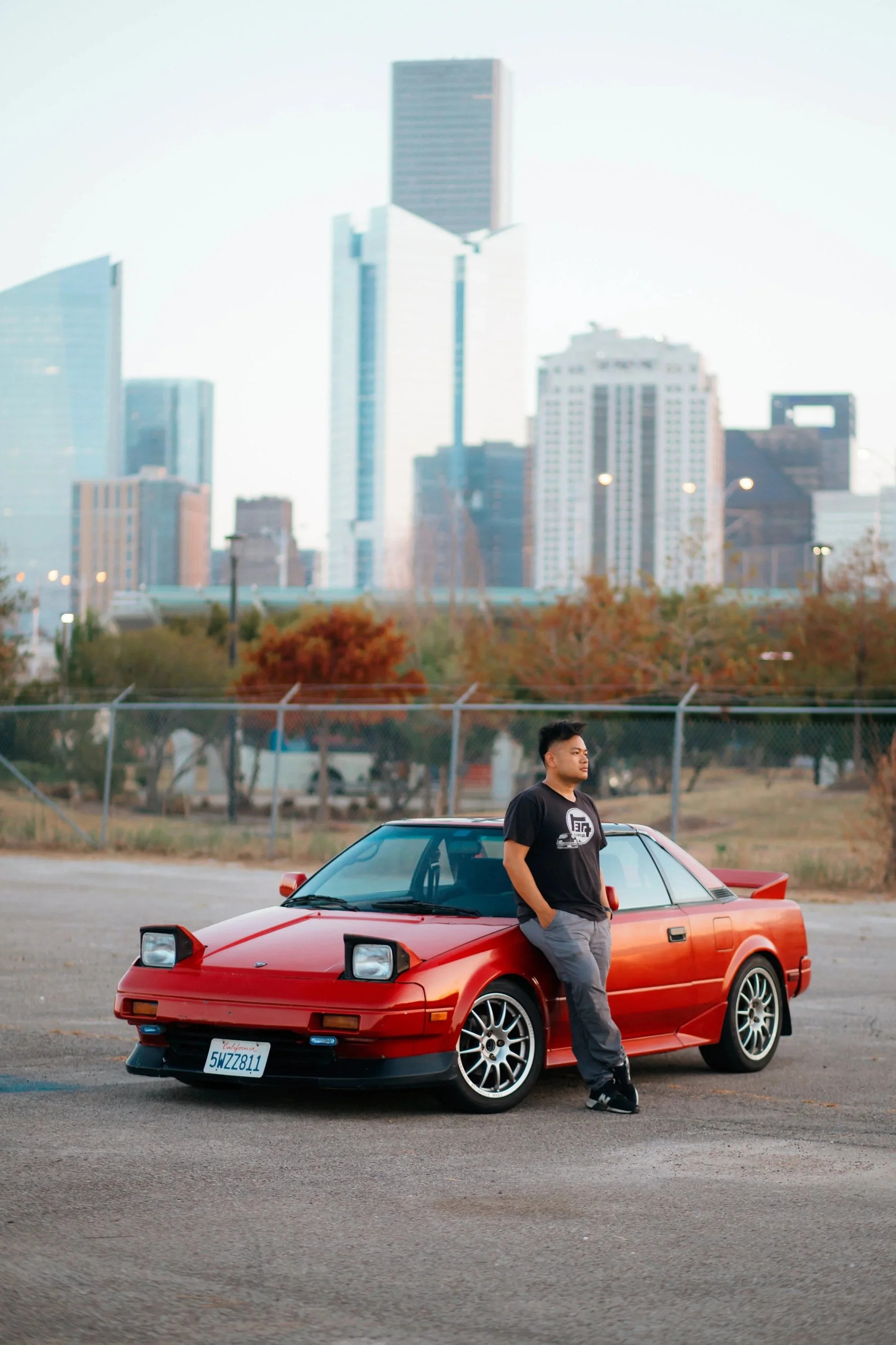 A young man standing next to a red sports car with his hands in pockets, outdoors in a parking lot with city skyscrapers in the background.