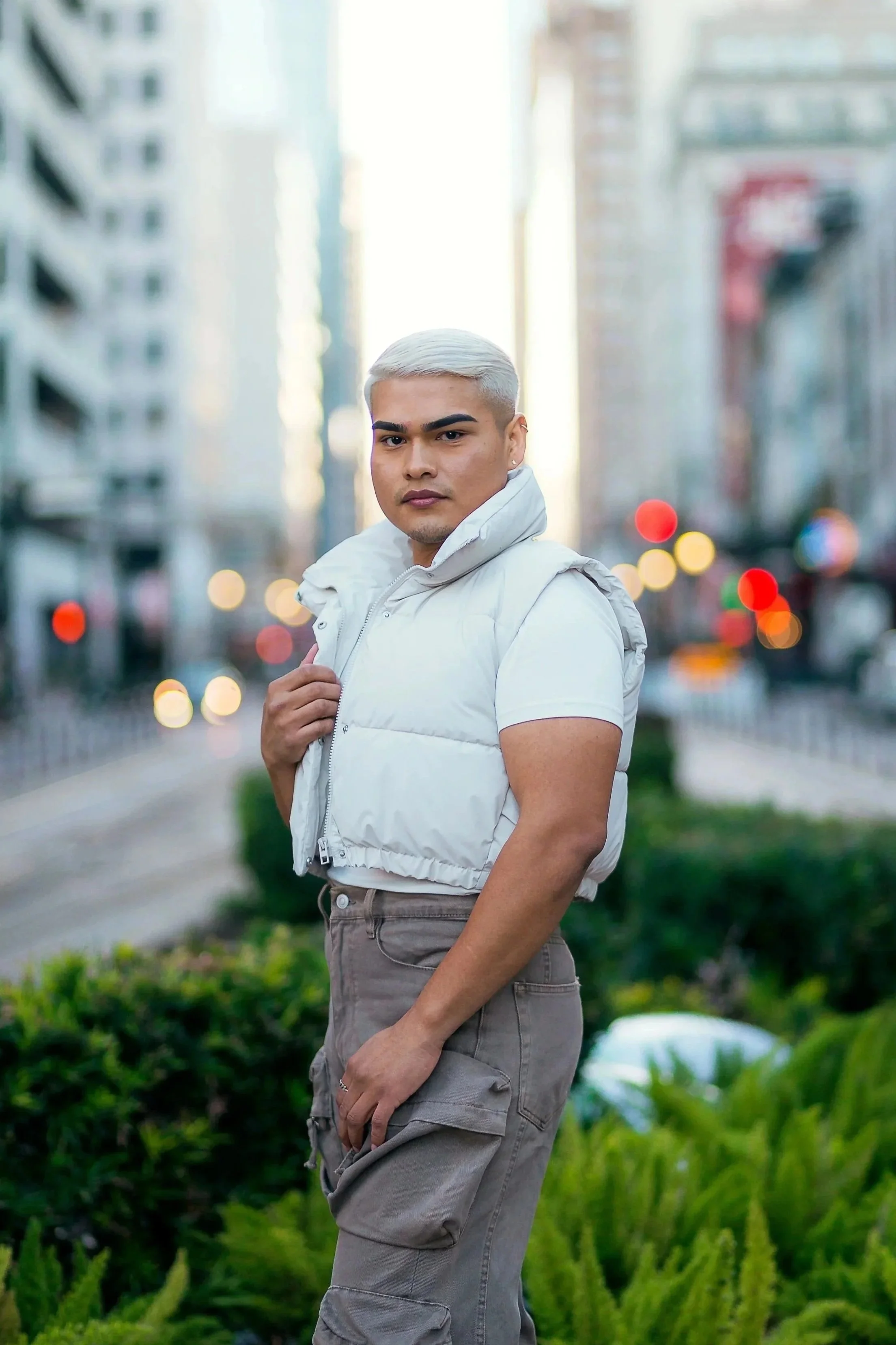 A man with platinum blonde hair wearing a white cropped vest over a white t-shirt standing outdoors in an urban setting with tall buildings and blurred lights in the background.