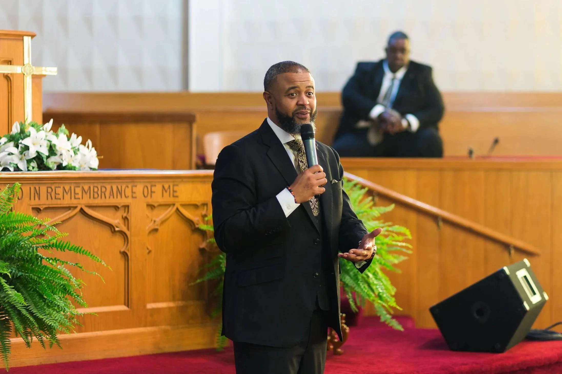 A man in a black suit holding a microphone speaking in a church. Another man sits in the background on a raised platform, with church decorations and greenery around.