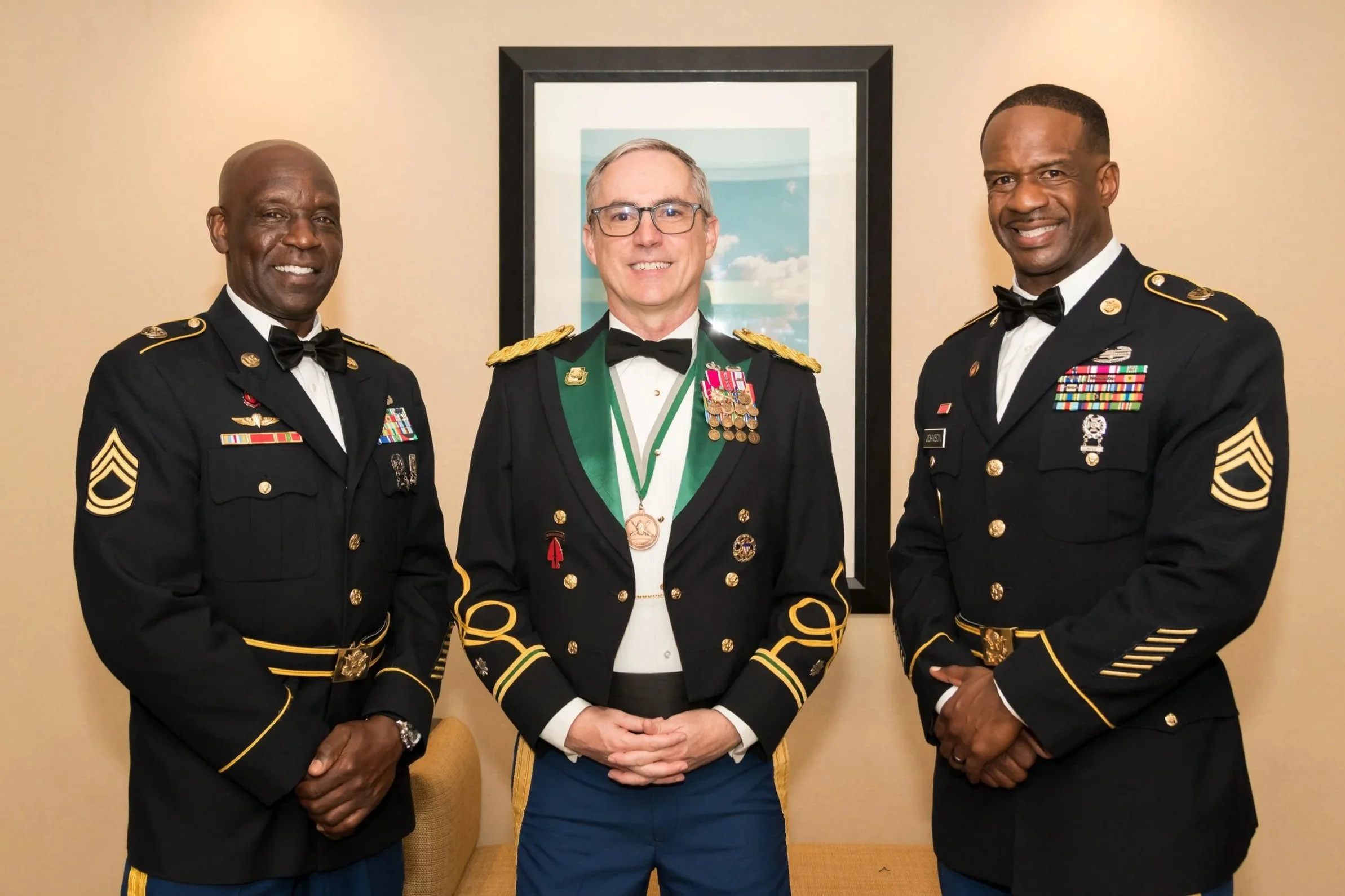 Three men in military dress uniforms standing indoors, smiling at the camera, with a framed picture of a cloudy sky behind them.