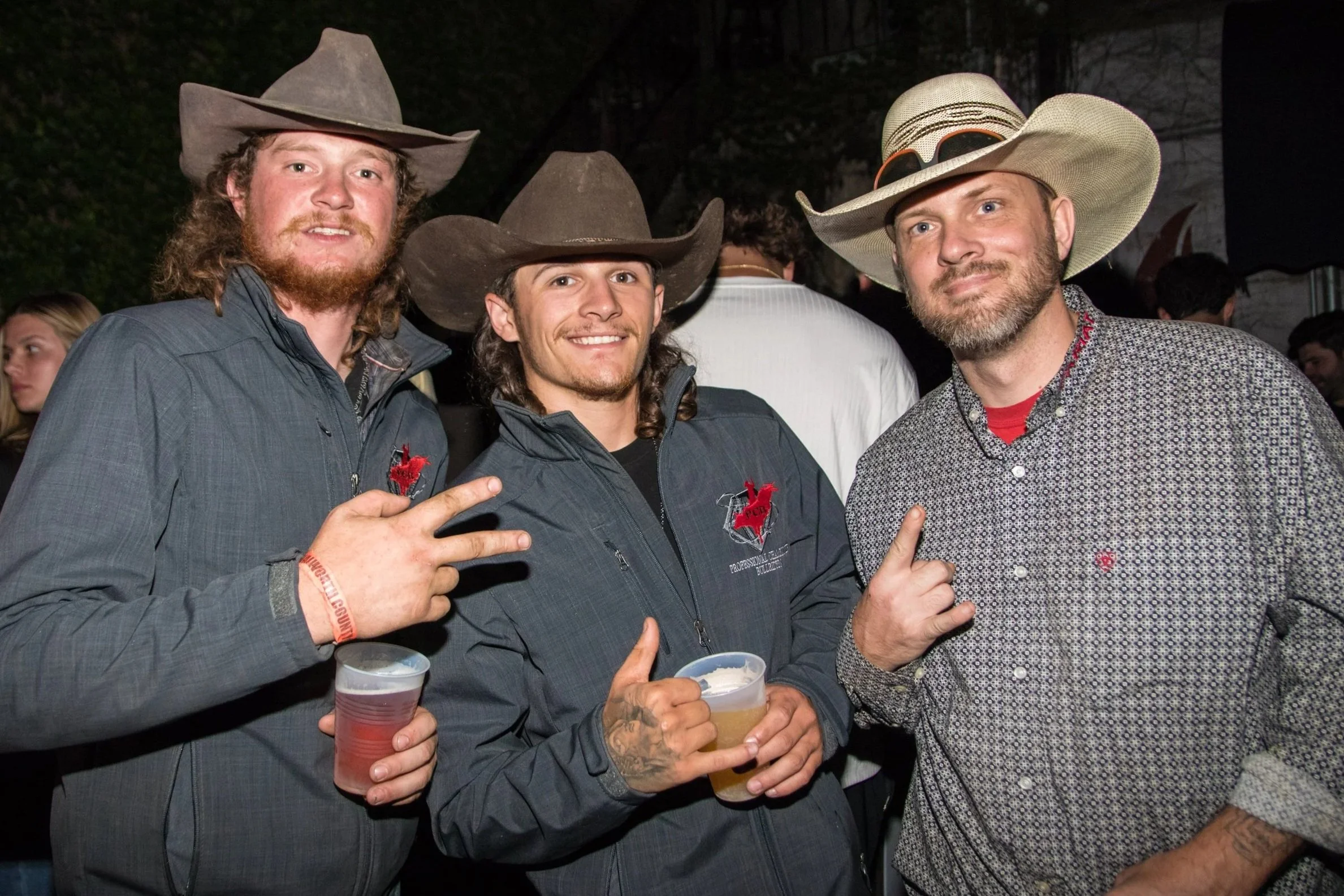 Three men wearing cowboy hats at an outdoor event, smiling, holding drinks, and making playful gestures.
