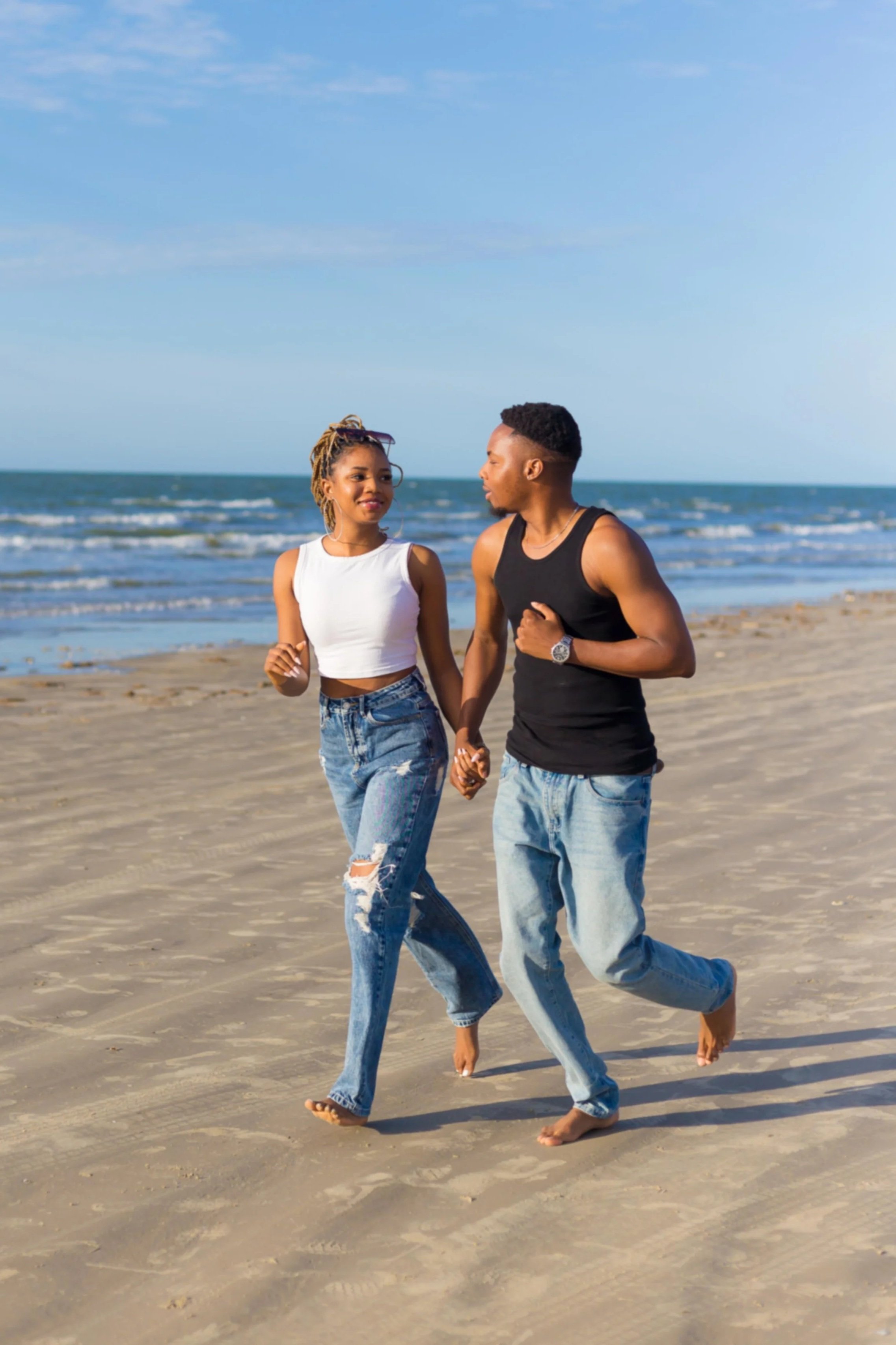 Soft romantic couple portrait by Levs Photography in Galveston, Texas featuring a young couple holding hands and walking barefoot along the beach near the ocean, smiling at each other under a bright blue sky with natural light and airy tones