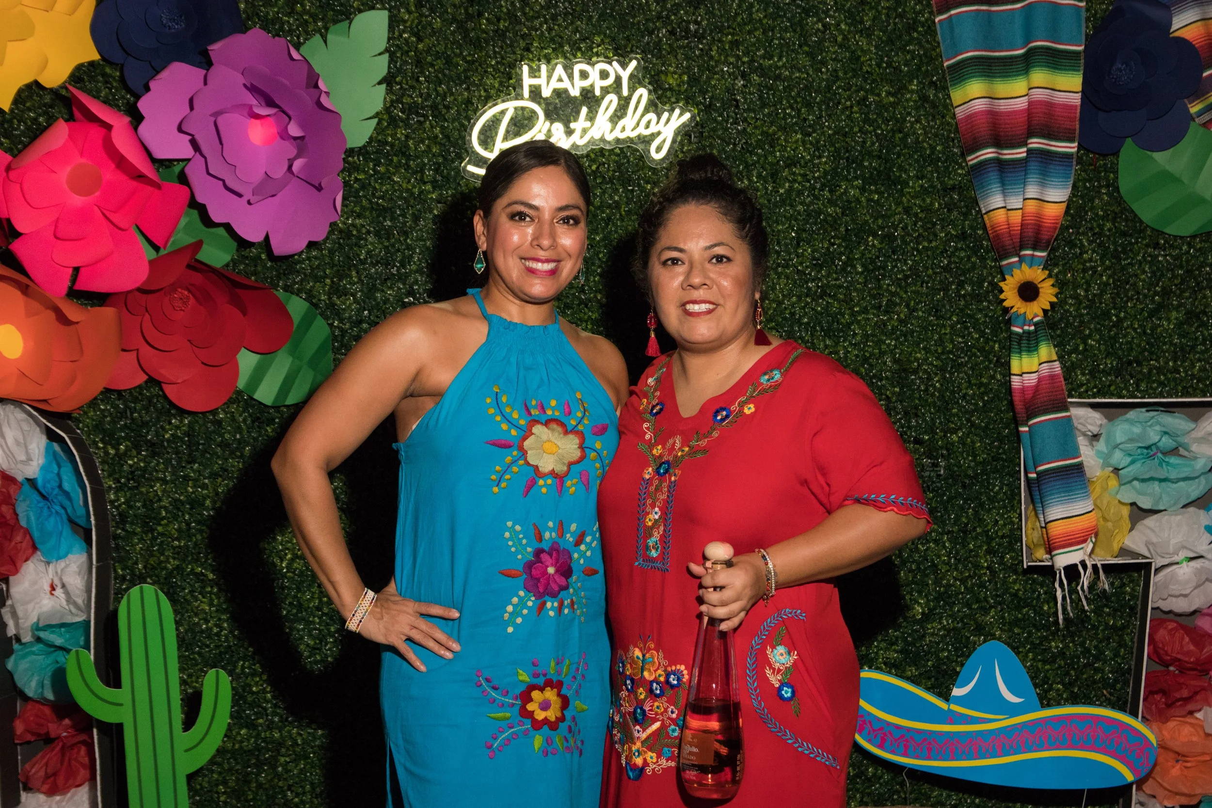 Two women smiling at a birthday party, standing in front of a green leafy backdrop with colorful paper flowers, a cactus, a hat, and a sign that reads 'Happy Birthday'.