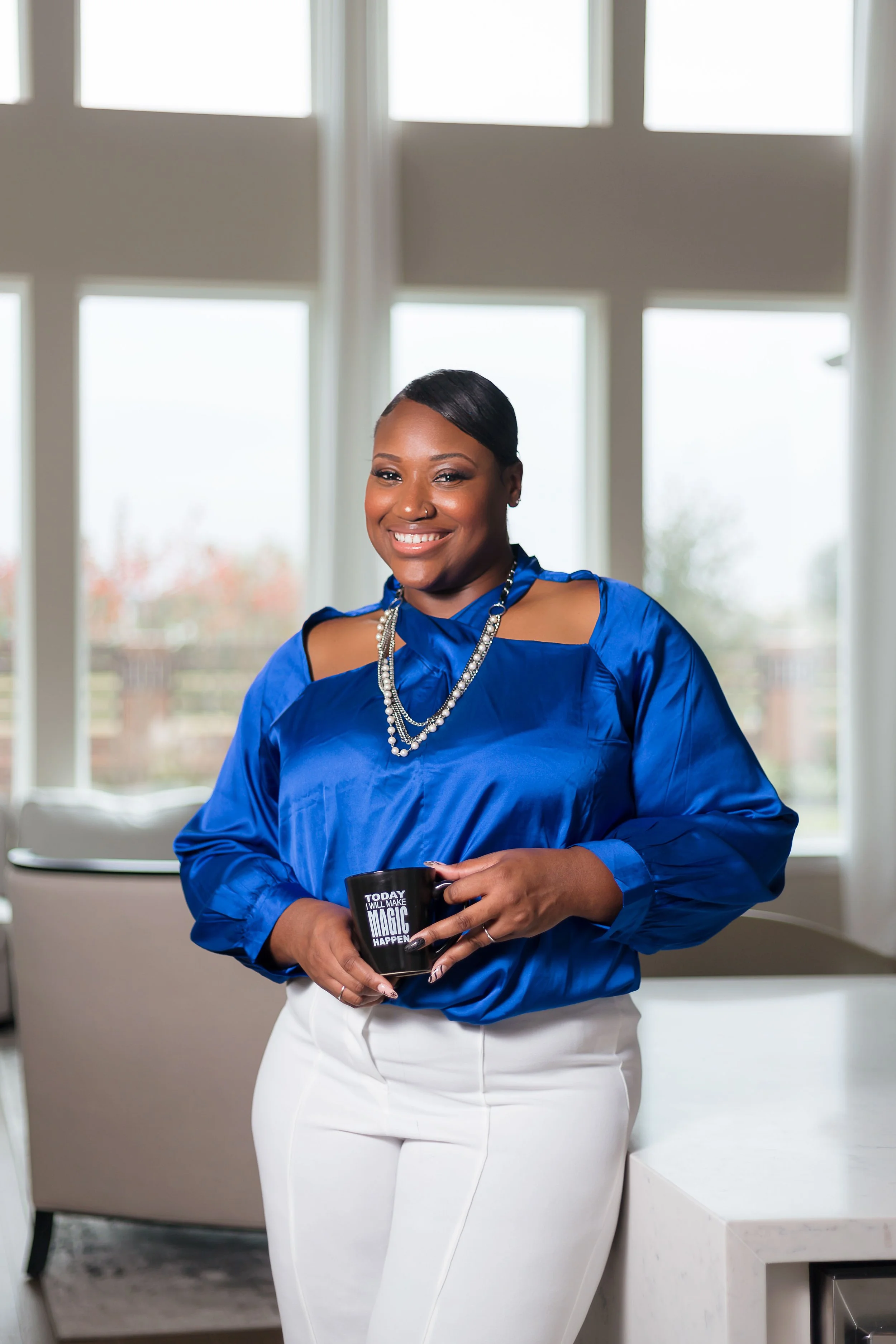 A woman with dark skin and short black hair smiling, wearing a shiny blue blouse with cut-out shoulders, white pants, and layered necklaces, holding a black coffee mug that says 'Today I Will Make Magic Happen,' standing in a bright room with large w