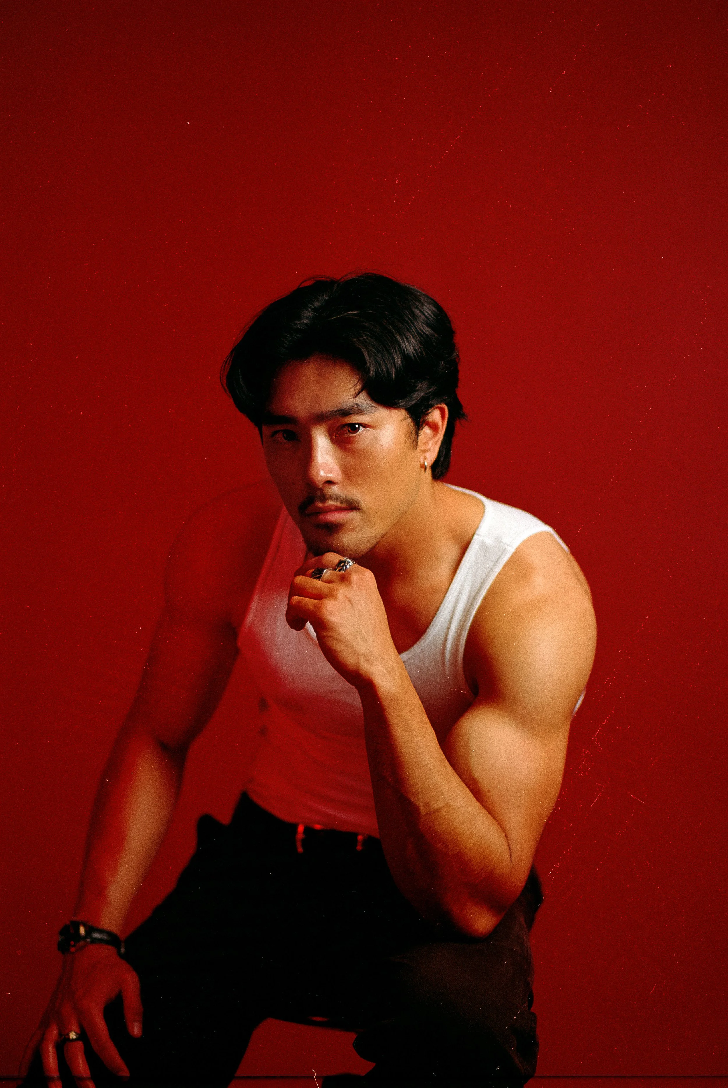 A young man with dark hair, wearing a white sleeveless shirt, is posing against a red background, with his chin resting on his hand.