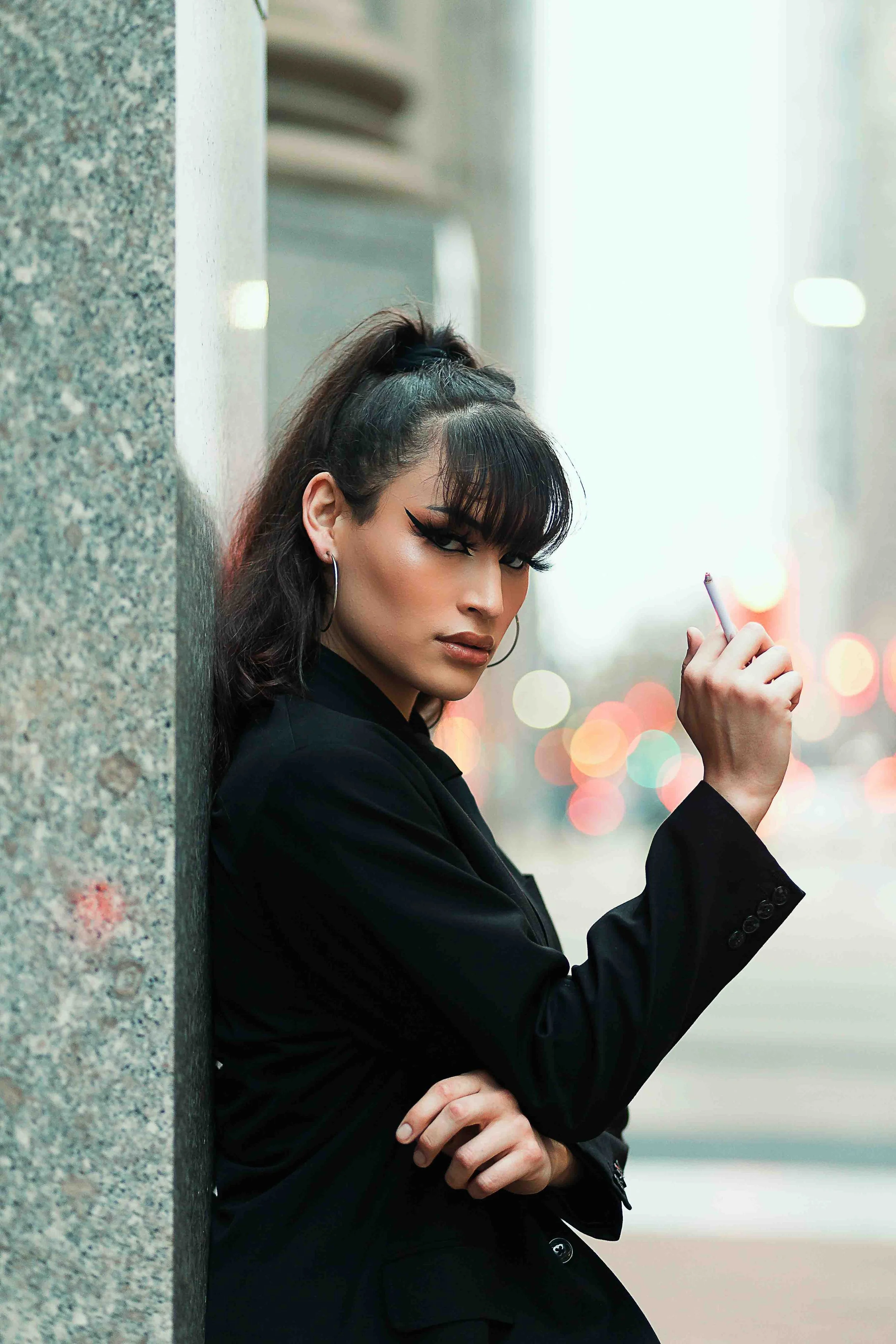 Downtown Houston editorial portrait by Lev's Photography featuring a woman with dark hair and makeup leaning against a concrete pillar, holding a cigarette on an urban street at dusk with blurred city lights, captured with boss-lady vibes for a cinem