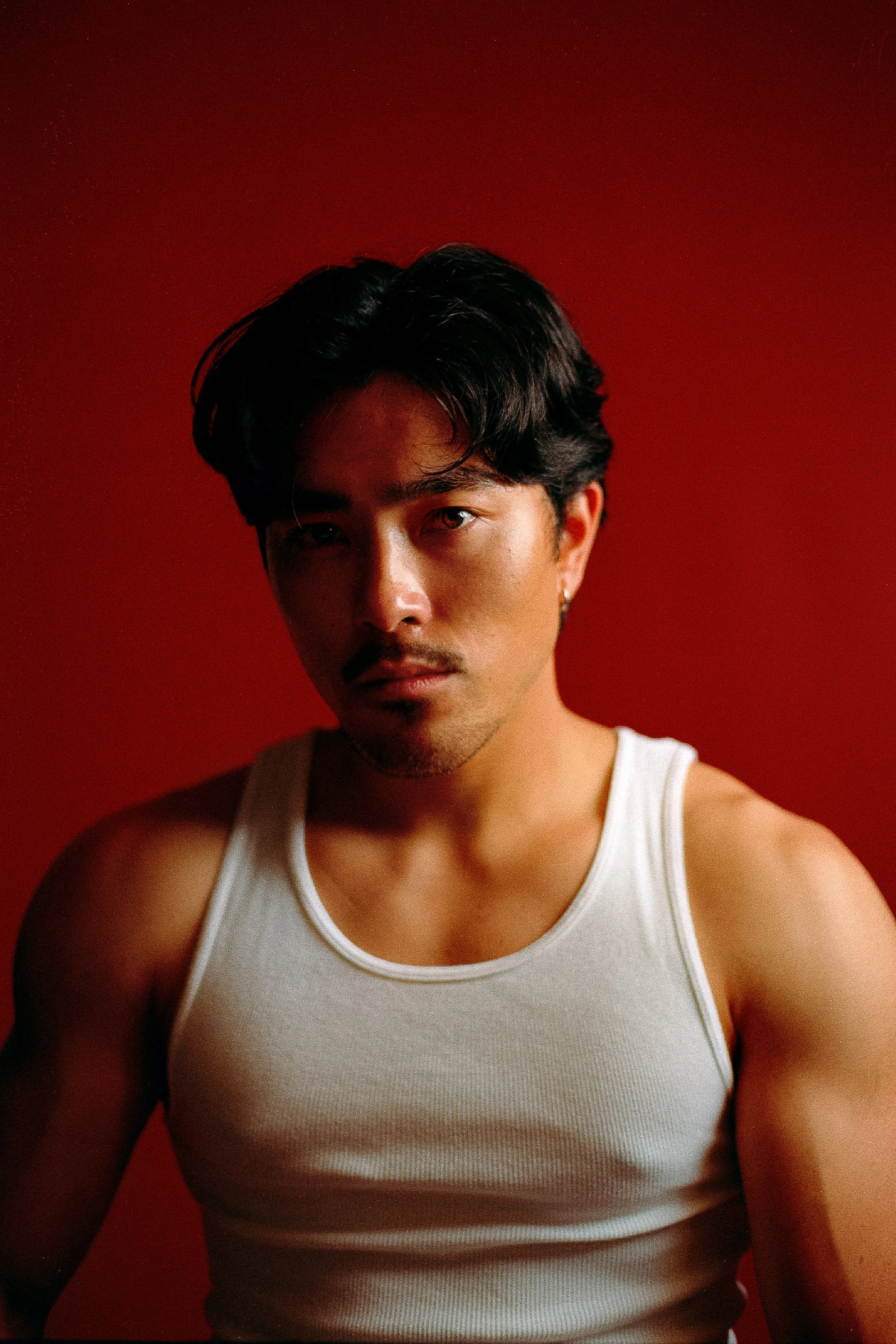 Portrait of a young man with dark hair, wearing a white tank top, against a red background.