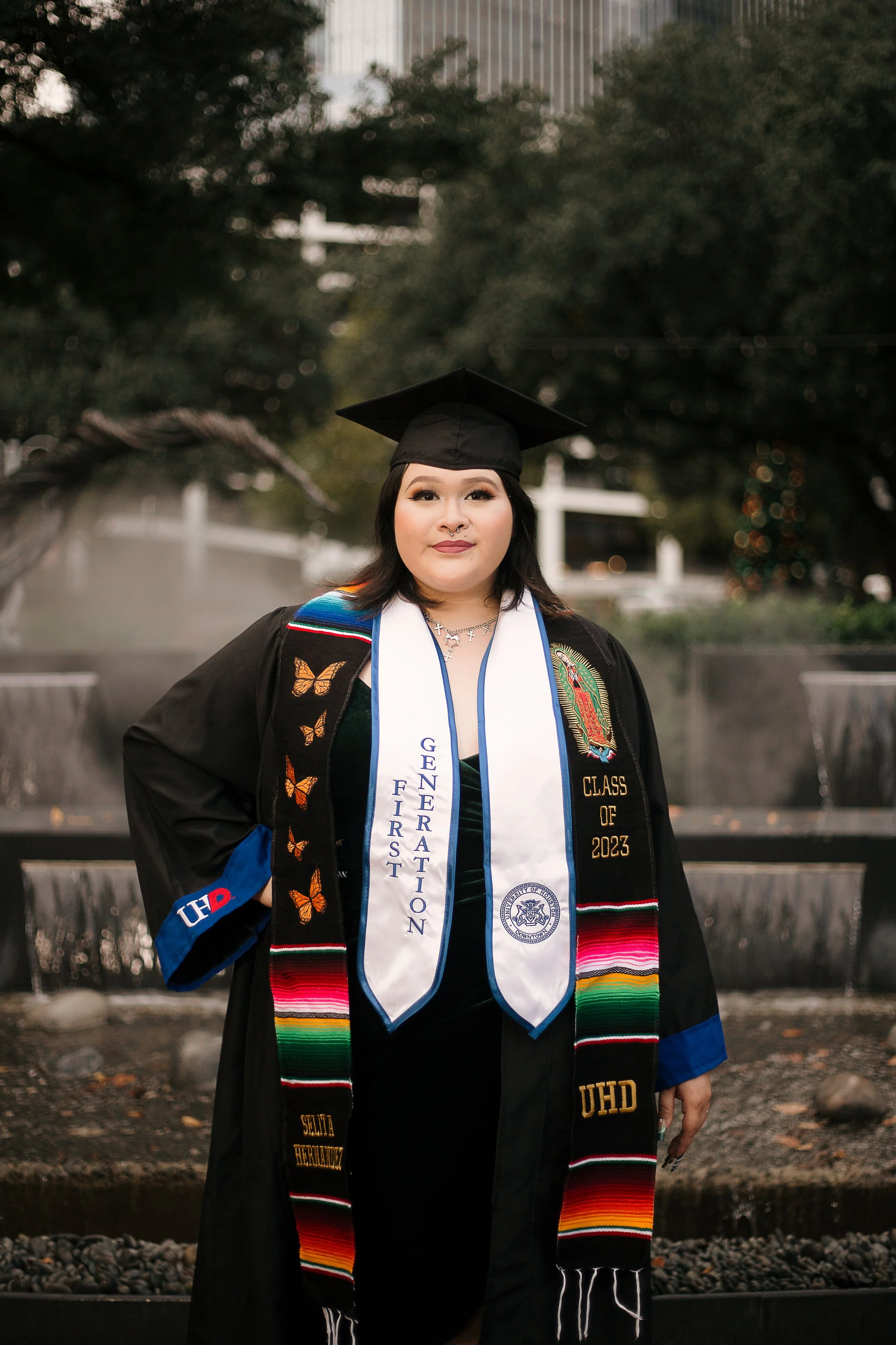 A young woman in a black graduation gown and cap stands outdoors in front of a fountain. She wears a white stole with 'GNERATlON FIRST' and the university seal, and a colorful sash with 'CLASS OF 2023' and 'UHD.' She has shoulder-length black hair, m
